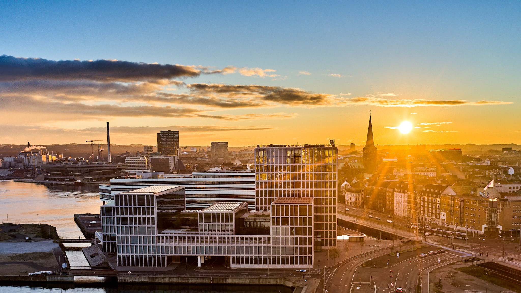 amex-magazin-aarhus-daenemark-hafenbucht In einer Hafenbucht steht ein modernes Gebäude im Sonnenschein.