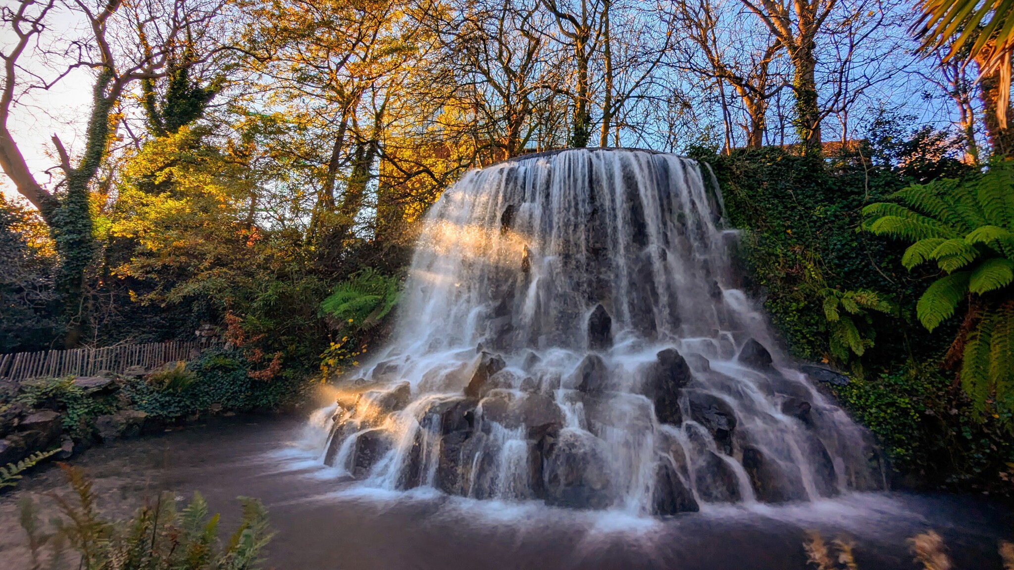 In einem Park stehen Steine, von denen ein Wasserfall in ein Becken fließt.