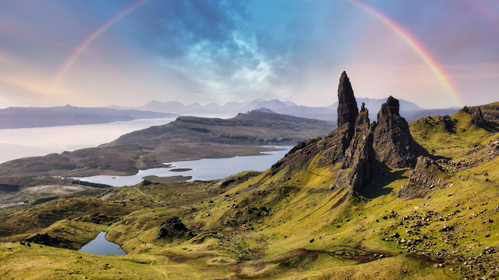 amex-magazin-isle-of-skye-regenbogen Drei spitze Felsen ragen auf einem Berg empor, im Hintergrund sind Flüsse mit einem Regenbogen zu sehen.