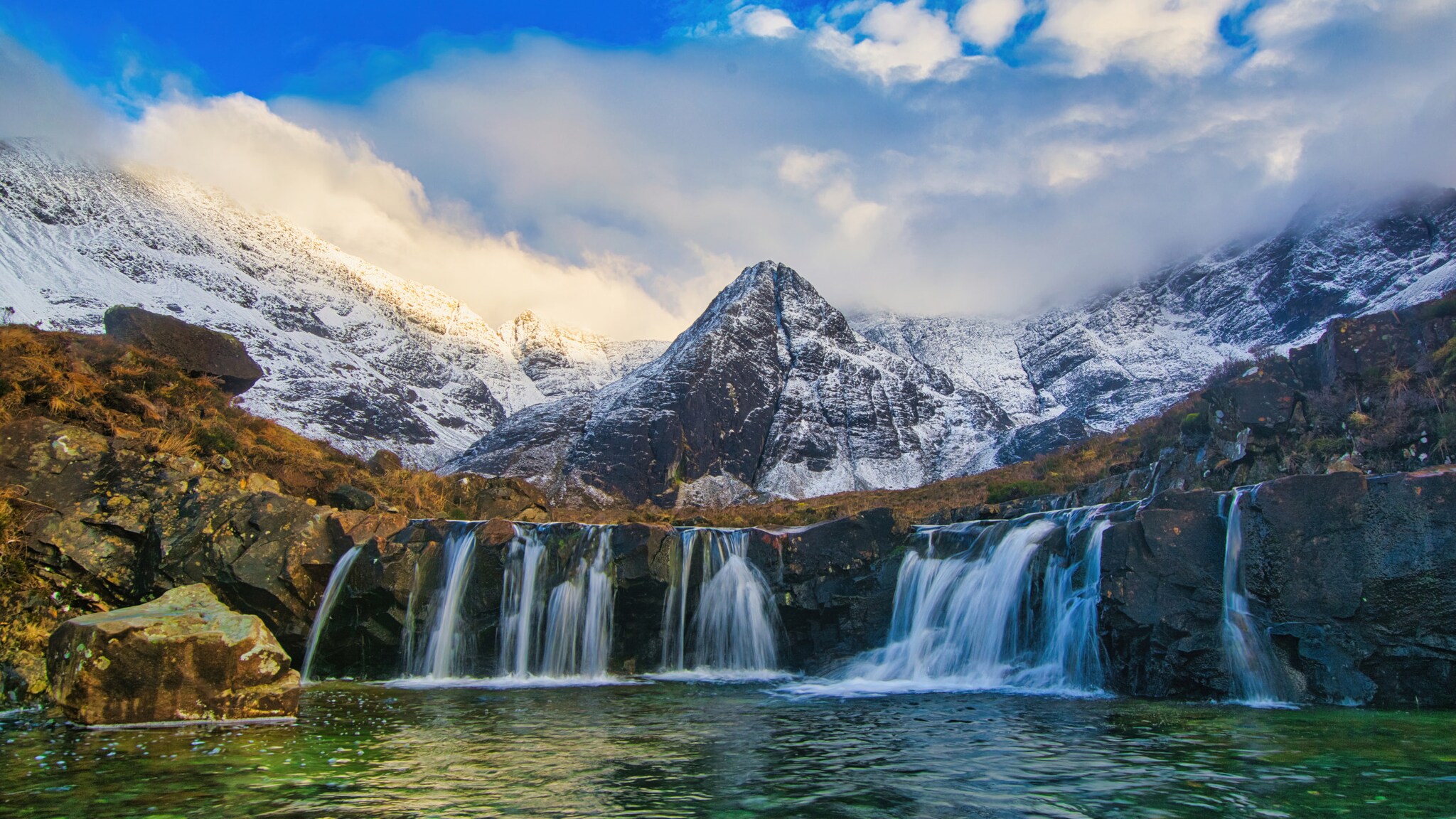 Kleine Wasserfälle fließen in ein Wasserbecken, im Hintergrund ragen Berge empor.