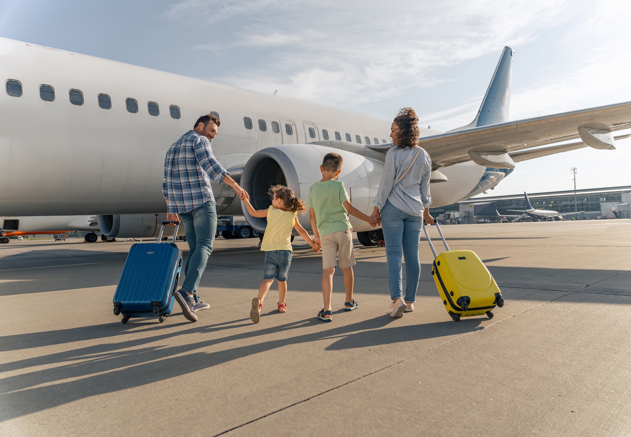 amex-insurances-reiseruecktritt-gruende-familie-vor-flugzeug Eine Familie läuft mit zwei Koffern über ein Rollfeld, ein Flugzeug ist im Hintergrund zu sehen.