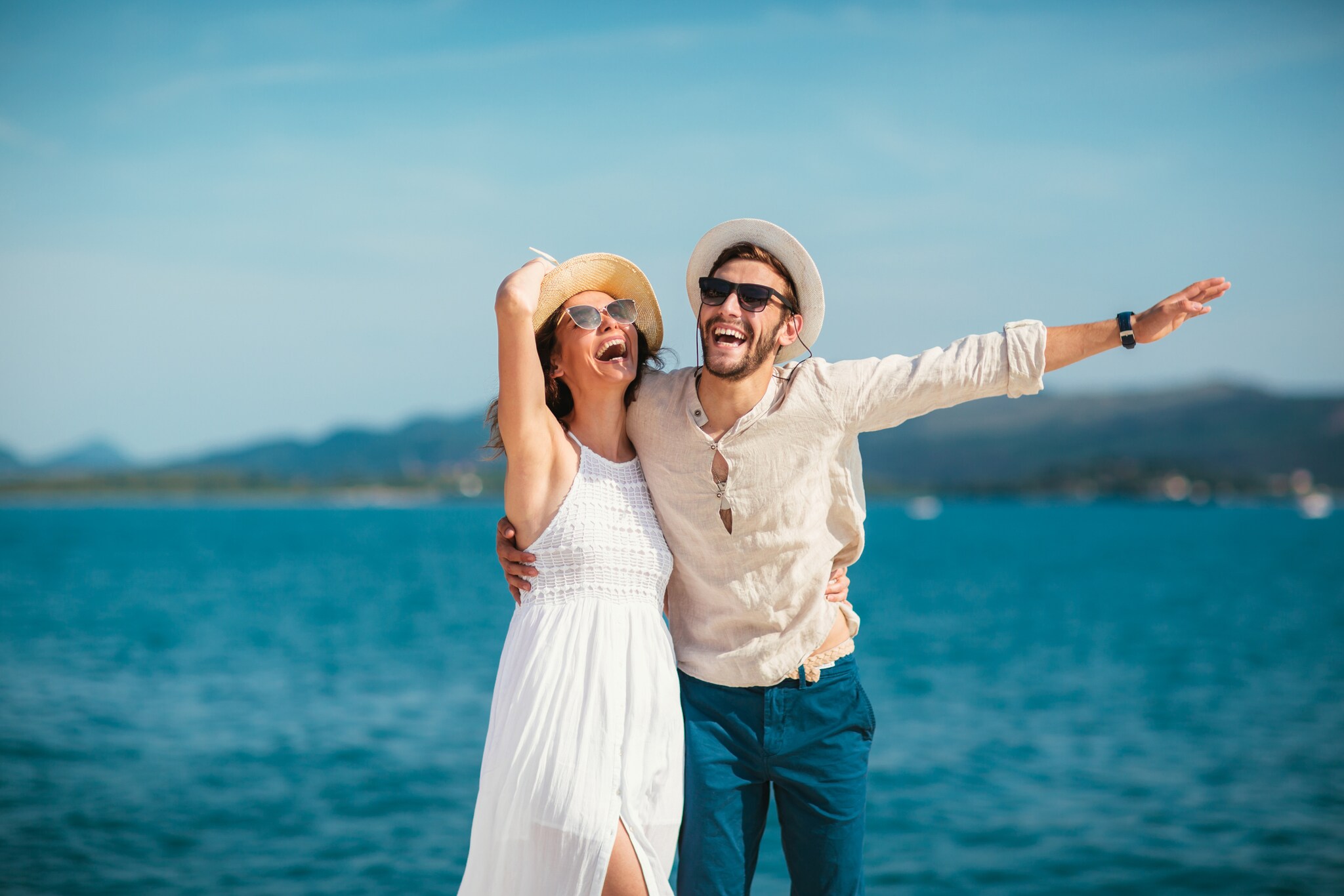 Ein Mann und eine Frau in sommerlichen Outfits stehen strahlend und lachend vor dem Meer.