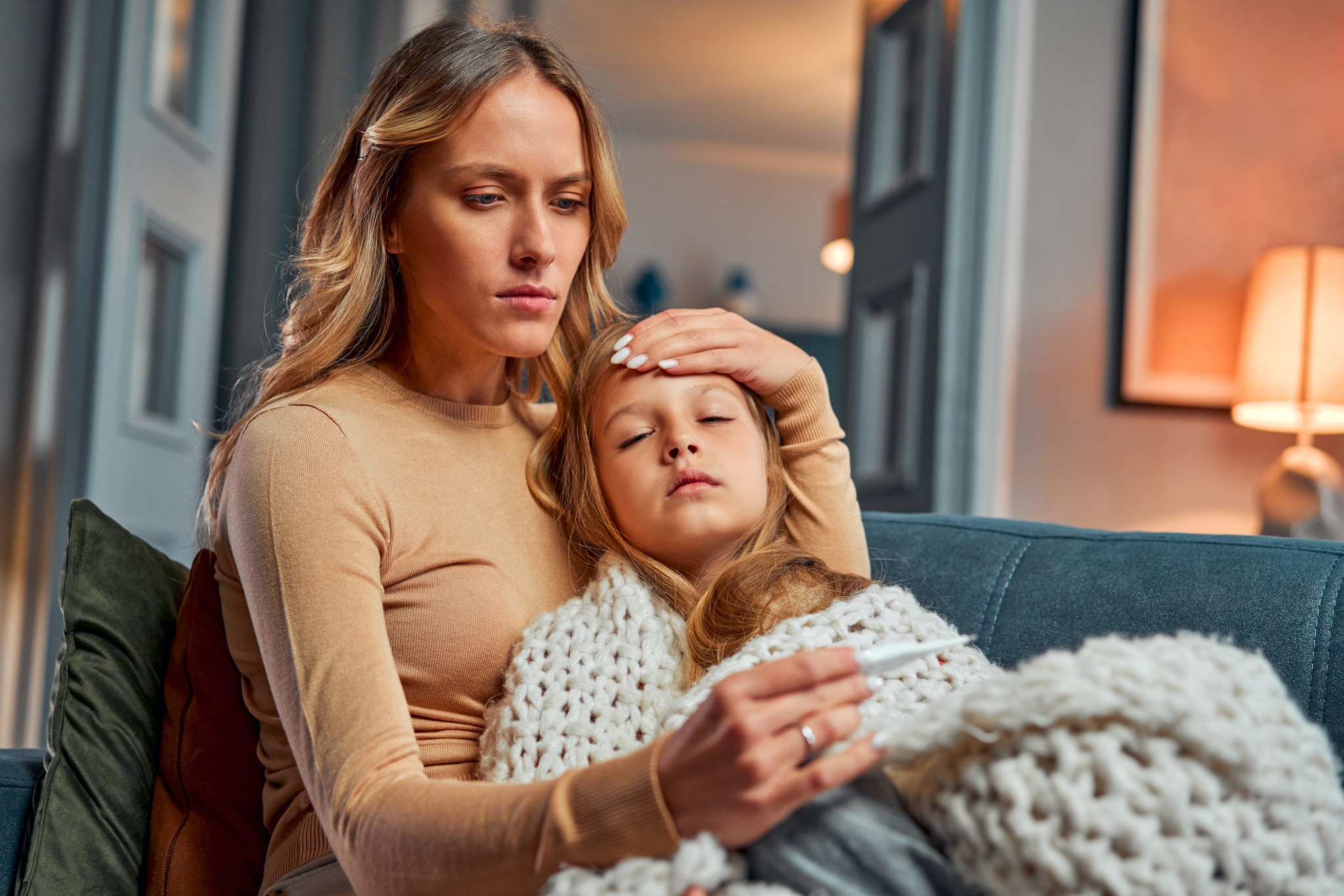 amex-insurances-reiseruecktritt-gruende-mutter-mit-krankem-kind Eine besorgt aussehende Mutter hält die Stirn ihrer Tochter, in der Hand hält sie ein Fieberthermometer.