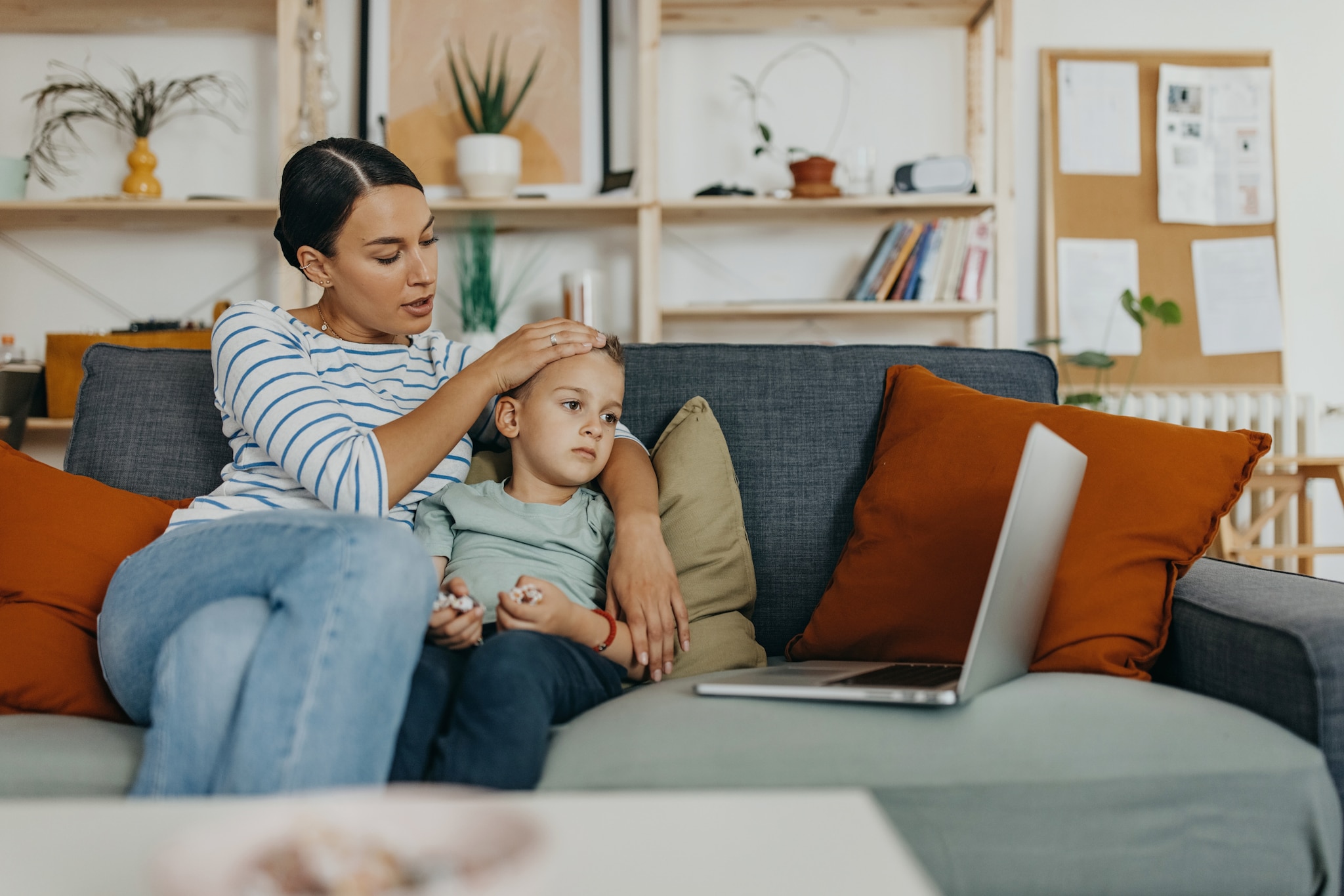 amex-insurances-reiseruecktritt-angehoerige-krank-mutter-mit-sohn Ein kleiner Junge sitzt auf dem Sofa, die Mutter hält ihre Hand an seine Stirn.