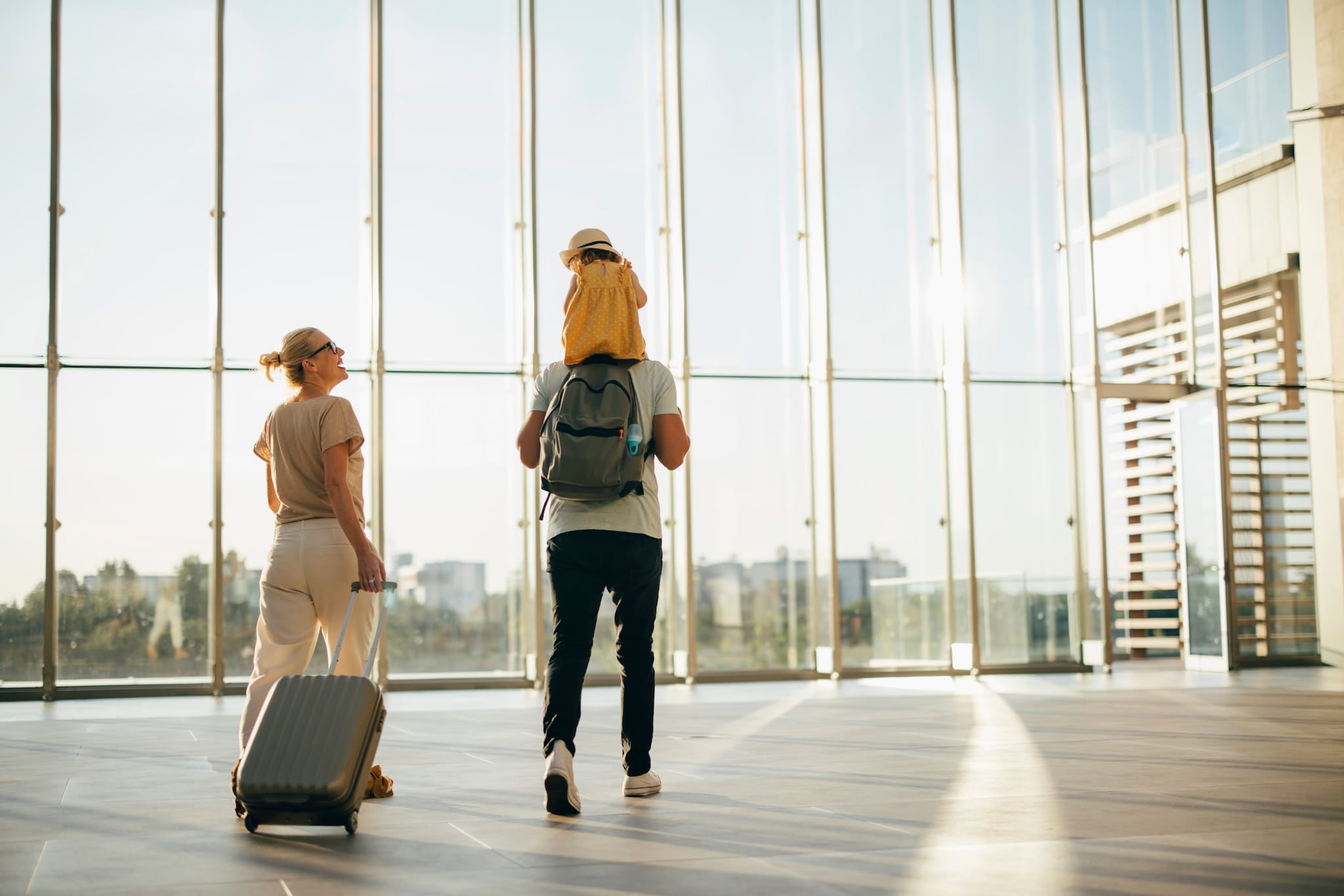 amex-insurances-reiseruecktritt-steuern-familie-flughafen Eine Frau, die einen Koffer zieht, lächelt ein Kind an, das auf den Schultern eines Mannes in einer Halle mit großen Glasfronten sitzt.