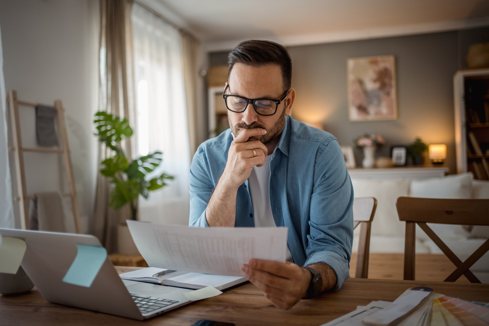 amex-insurances-reiseruecktritt-steuern-mann-laptop Ein Mann, der an einem Tisch vor einem Laptop sitzt und in ein Dokument schaut, das er in der Hand hält.