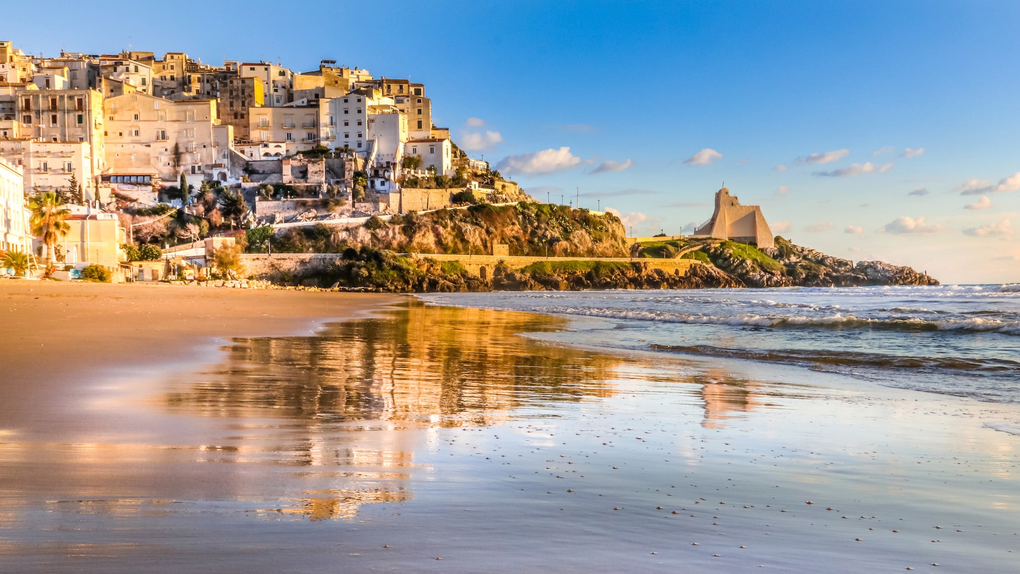 amex-magazin-rom-urlaub-strand-felsen Hinter einem weiten Strand ragen Gebäude und Felsen empor.