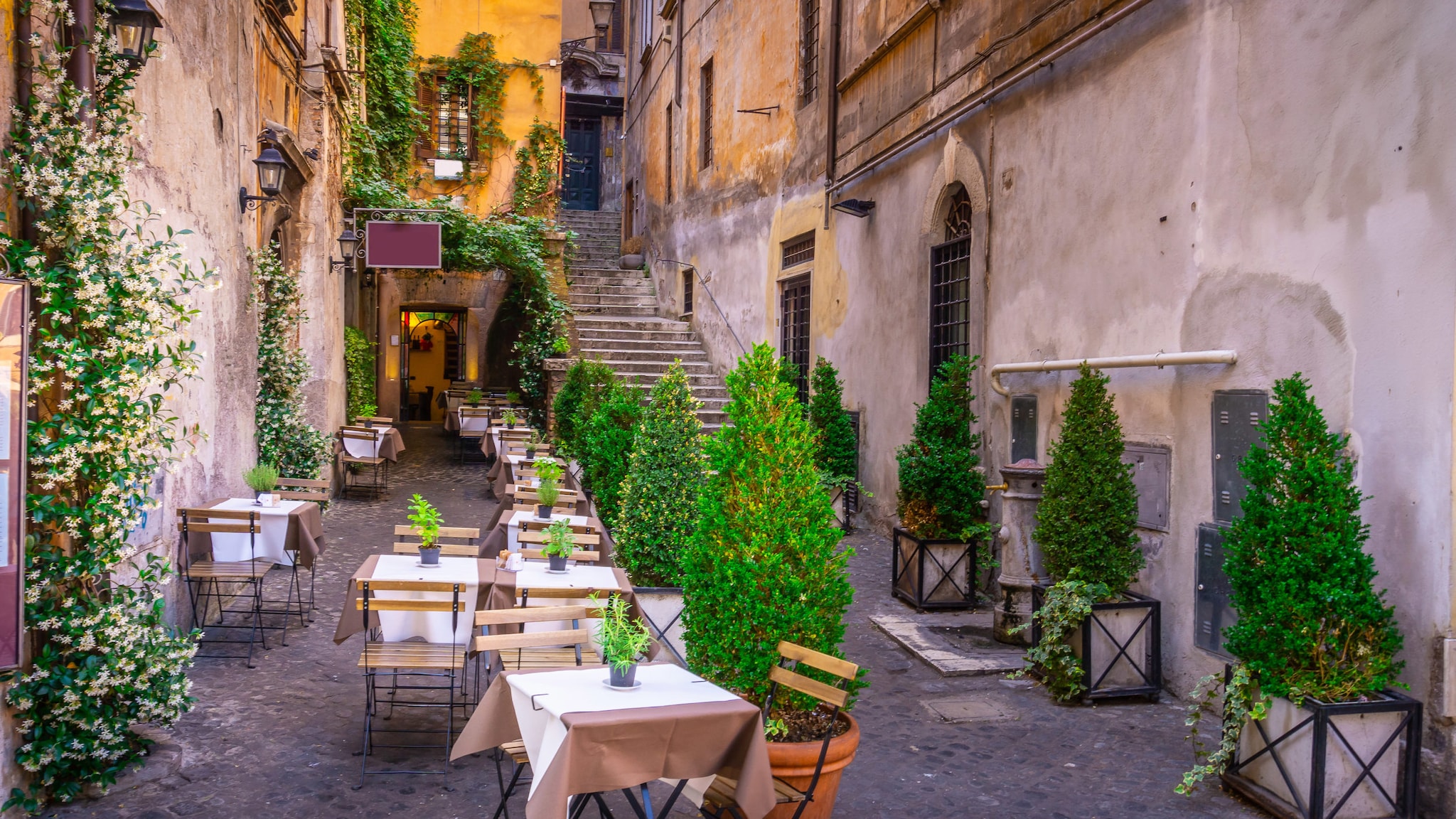 amex-magazin-rom-urlaub-treppe-innenhof In einem kleinen Innenhof mit alter Treppe stehen rustikale Tische mit Stühlen.