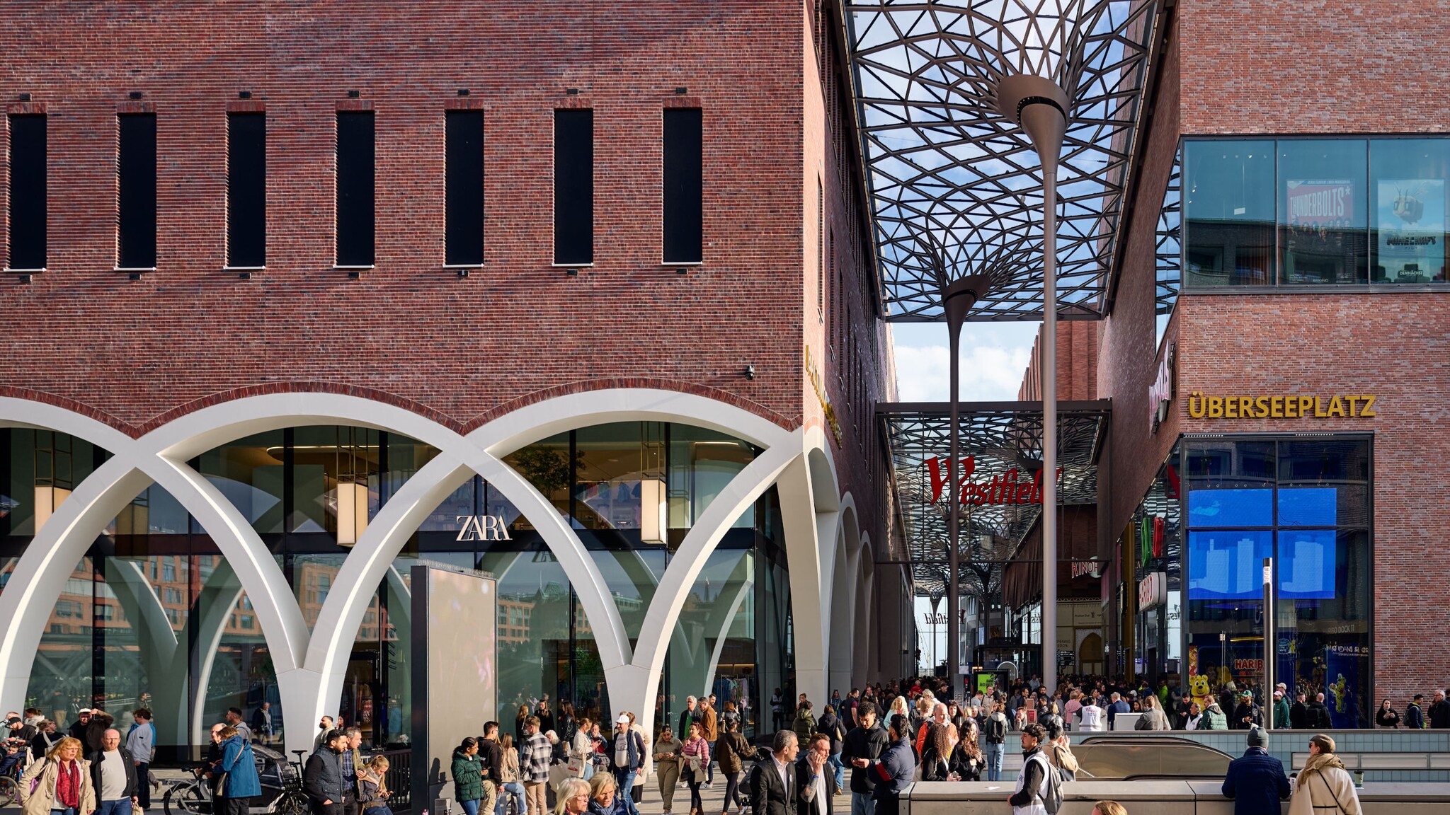 Personen laufen in einem Einkaufszentrum mit Backsteinfassade und Glasarchitektur unter freiem Himmel.