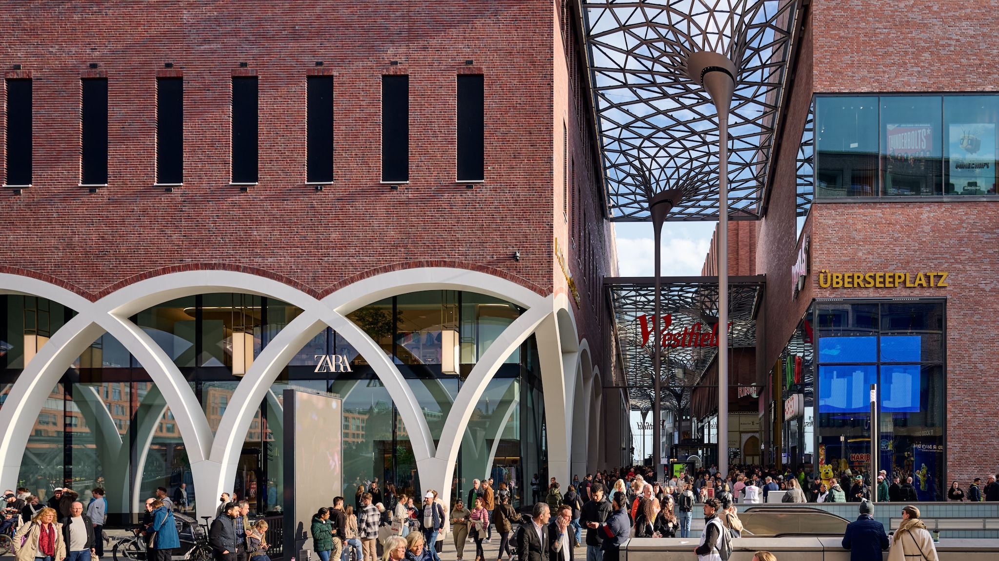 amex-magazin-westfield-hamburg-einkaufszentrum-backsteinfassade-Daniel_Schaefer Personen laufen in einem Einkaufszentrum mit Backsteinfassade und Glasarchitektur unter freiem Himmel.