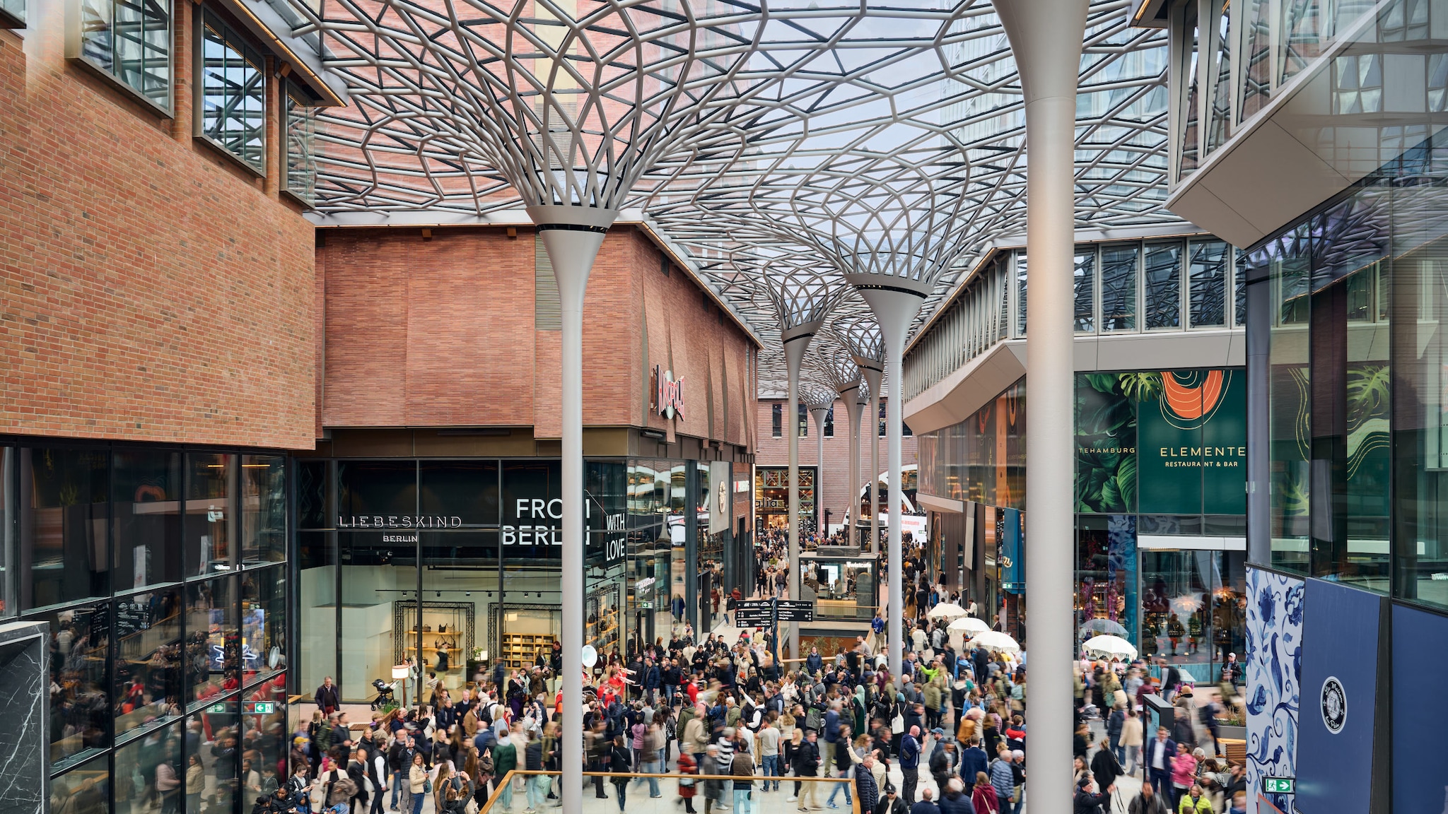 amex-magazin-westfield-hamburg-einkaufszentrum-laeden-Daniel_Schäfer Personen laufen durch ein halboffenes Einkaufszentrum mit einem organisch geformten Dach aus einer Glas- und Stahlkonstruktion.