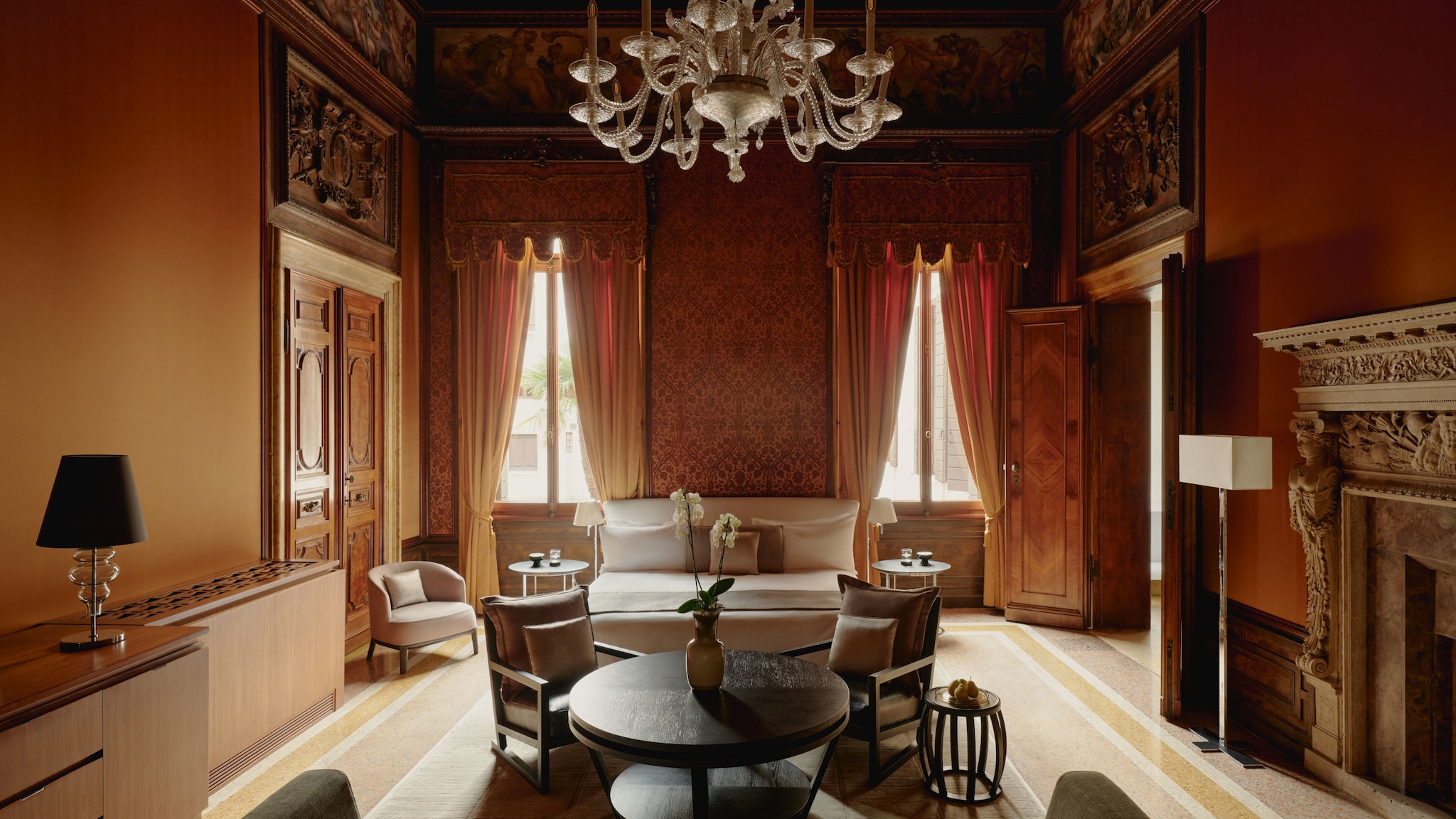 amex-magazin-venedig-urlaub-amanvenice-bedroom In einem hohen Raum mit verzierten Türen und Kamin steht ein Tisch mit Stühlen vor zwei großen Fenstern mit Vorhängen.