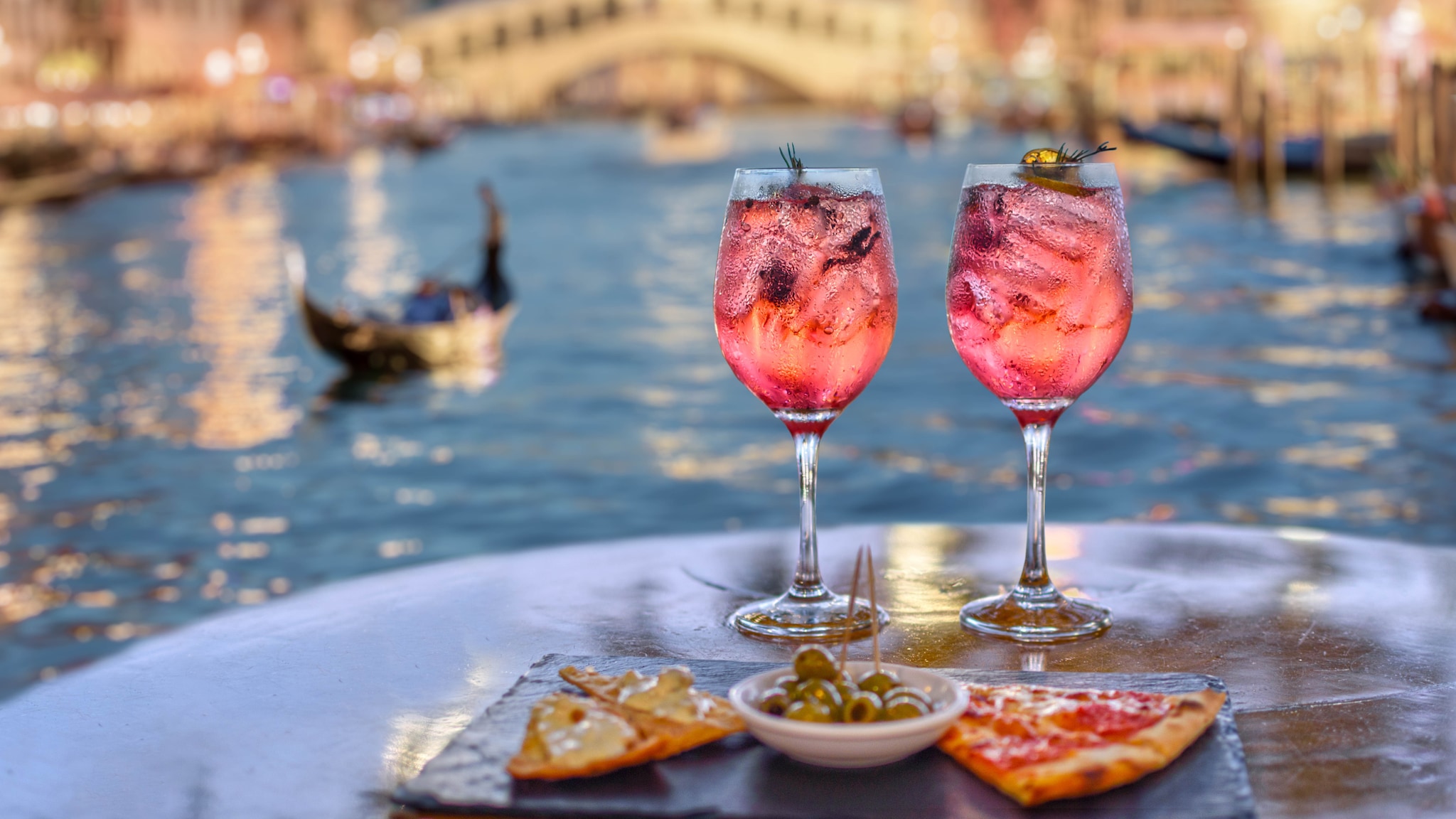 amex-magazin-venedig-urlaub-glaeser-wein Zwei Gläser Wein und Tapas stehen auf einem Tisch, im Hintergrund ist ein Fluss mit kleiner Brücke.