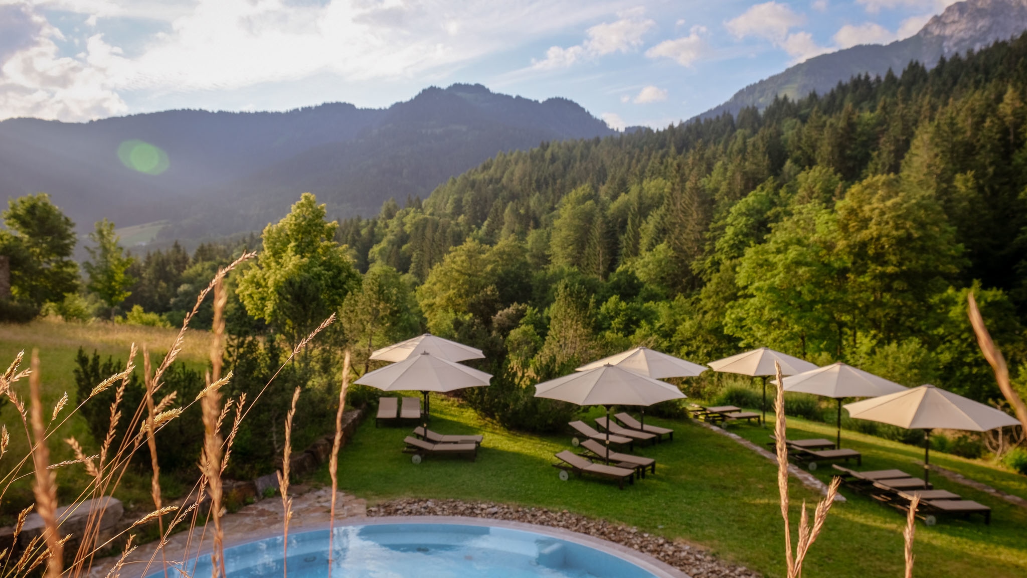 amex-magazin-urlaub-in-den-bergen-pool Sonnenliegen mit Schirmen auf einer Rasenfläche an einem Pool vor bewaldeter Berglandschaft im Sommer.