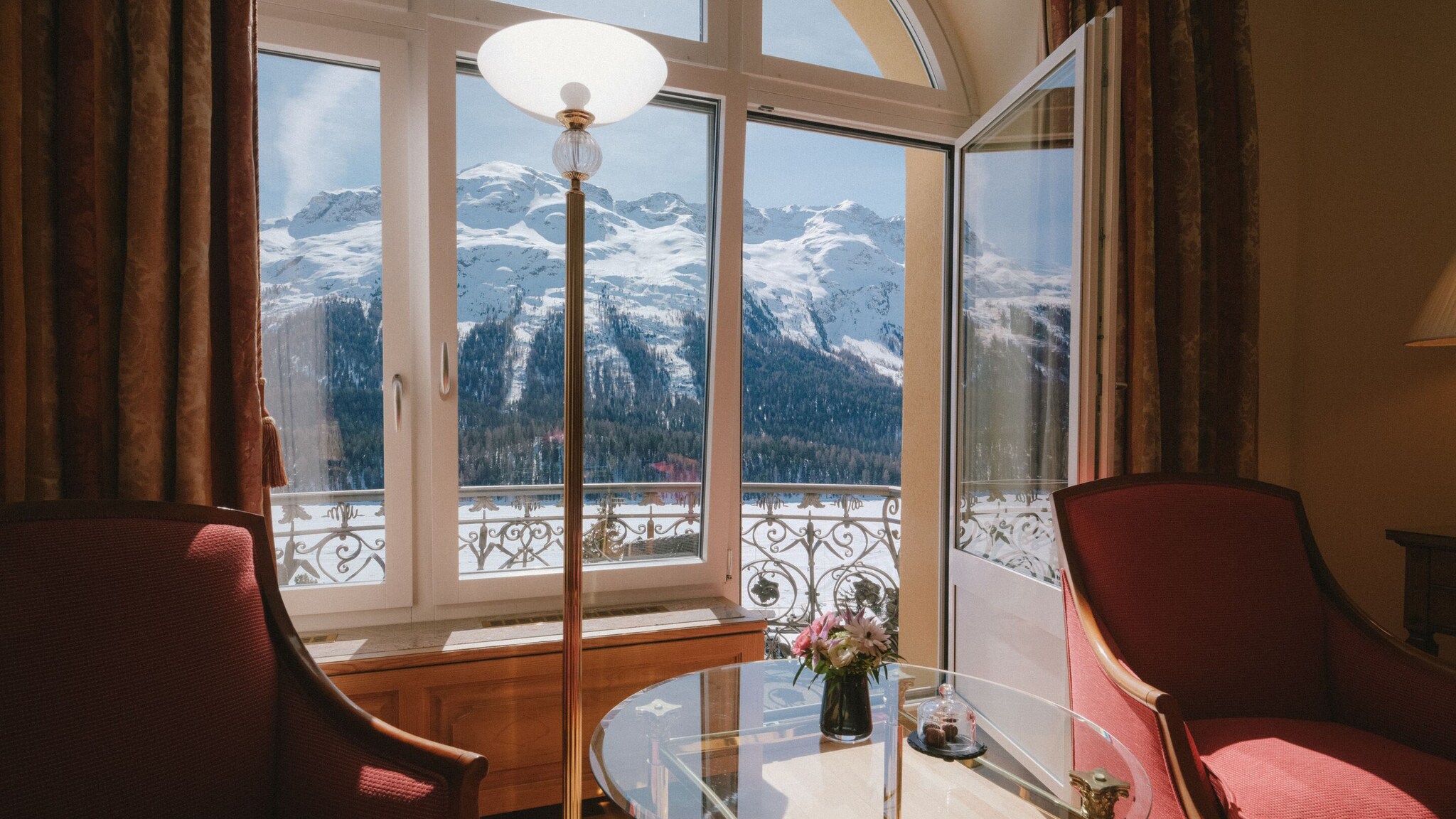 Ausblick aus einem Zimmer mit zwei Sesseln vor einer geöffneten Balkontür auf eine verschneite Berglandschaft.