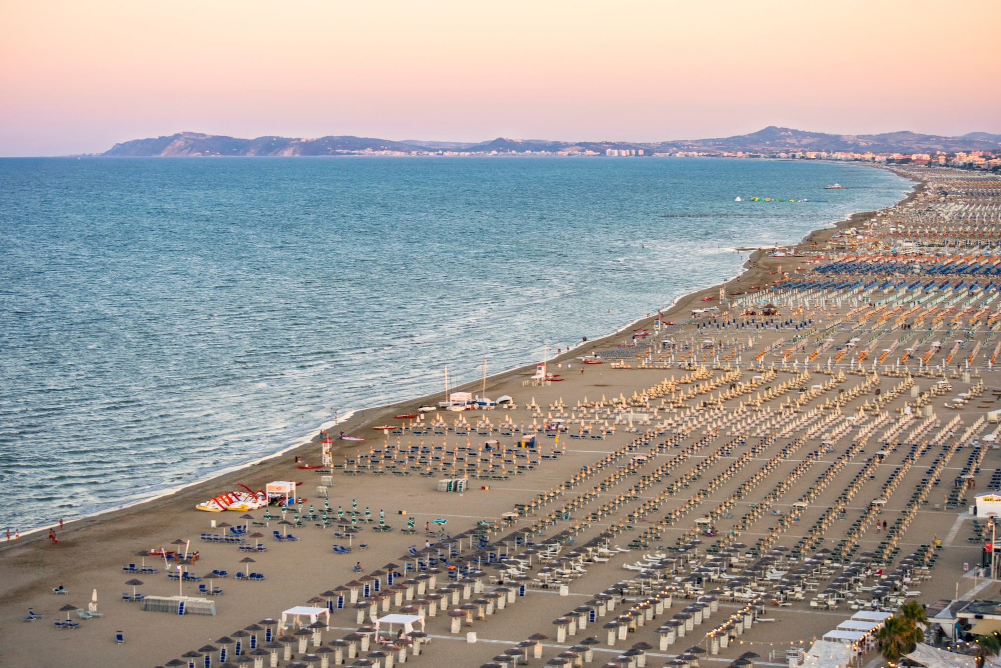 amex-magazin-rimini-italien-strand-meer Riminis breiter und langer Sandstrand von oben, mit bunten Sonnenschirmen, dem tiefblauen Meer und einem rosa gefärbten Himmel.