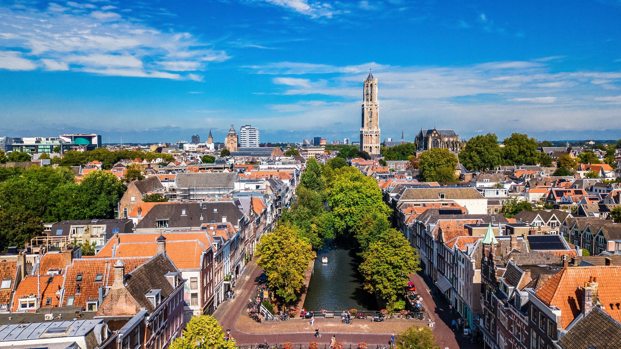 amex-magazin-utrecht-niederlanden-kanal-stadt Luftaufnahme einer Stadt mit Kanal, Bäumen und einem Domturm, der die umliegenden Häuser überragt.