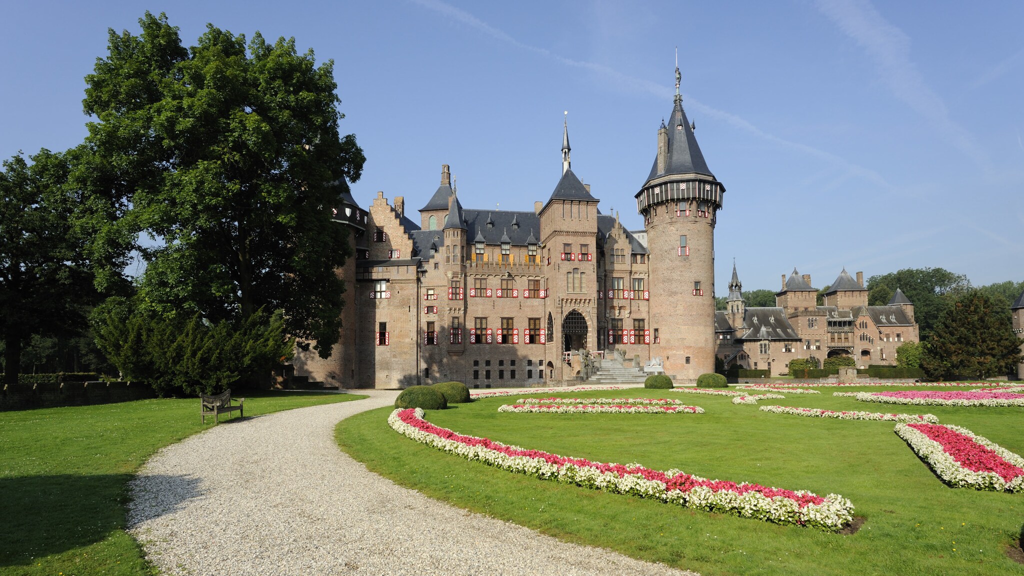 Ein historisches Schloss mit Turm, zu dem ein Schotterweg führt, umgeben von einer Parkanlage mit Blumenbeeten, Wiesen und Bäumen.
