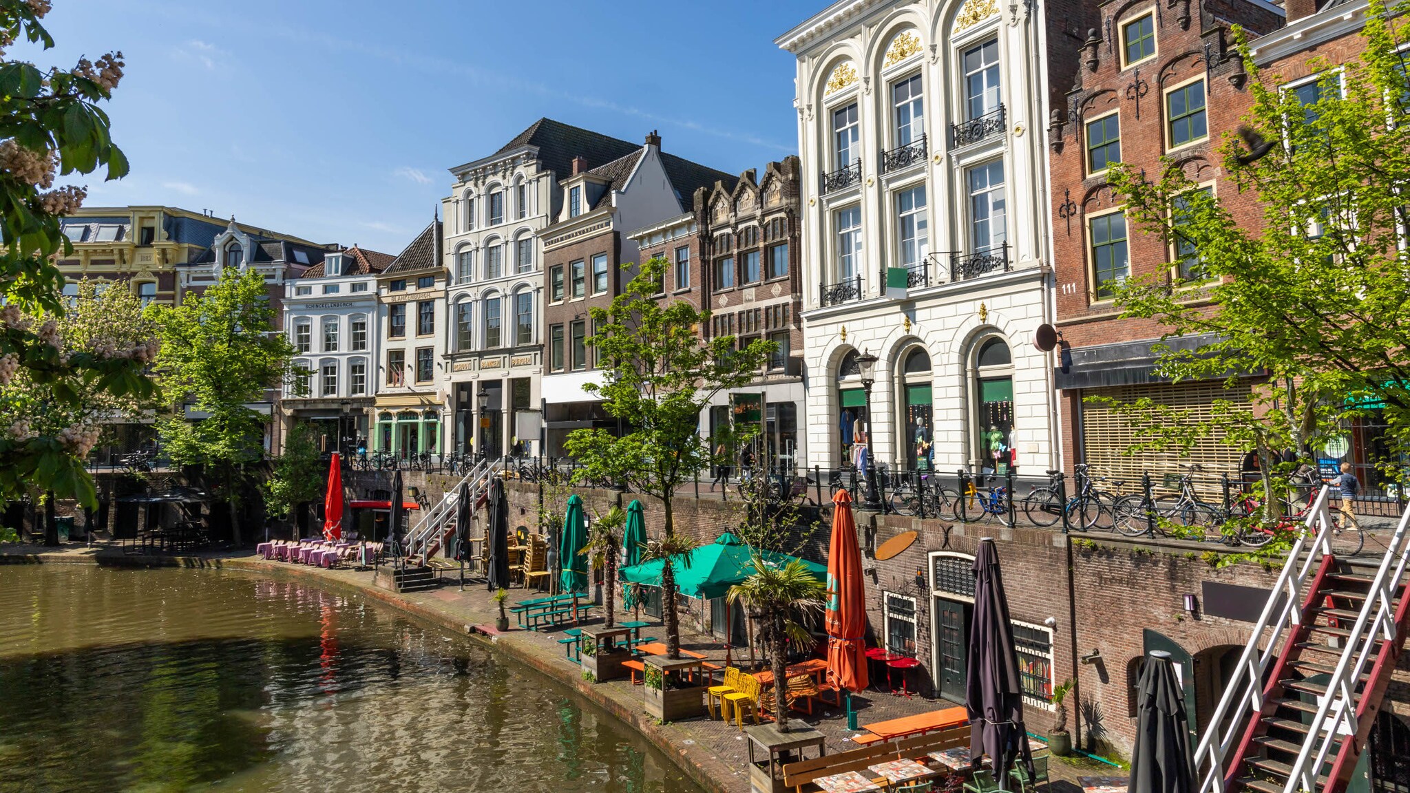 Restaurants mit Tischen, Bänken und Sonnenschirmen am Ufer eines Kanals, darüber liegt eine weitere Ebene mit Promenade und bunten Häuserfassaden.