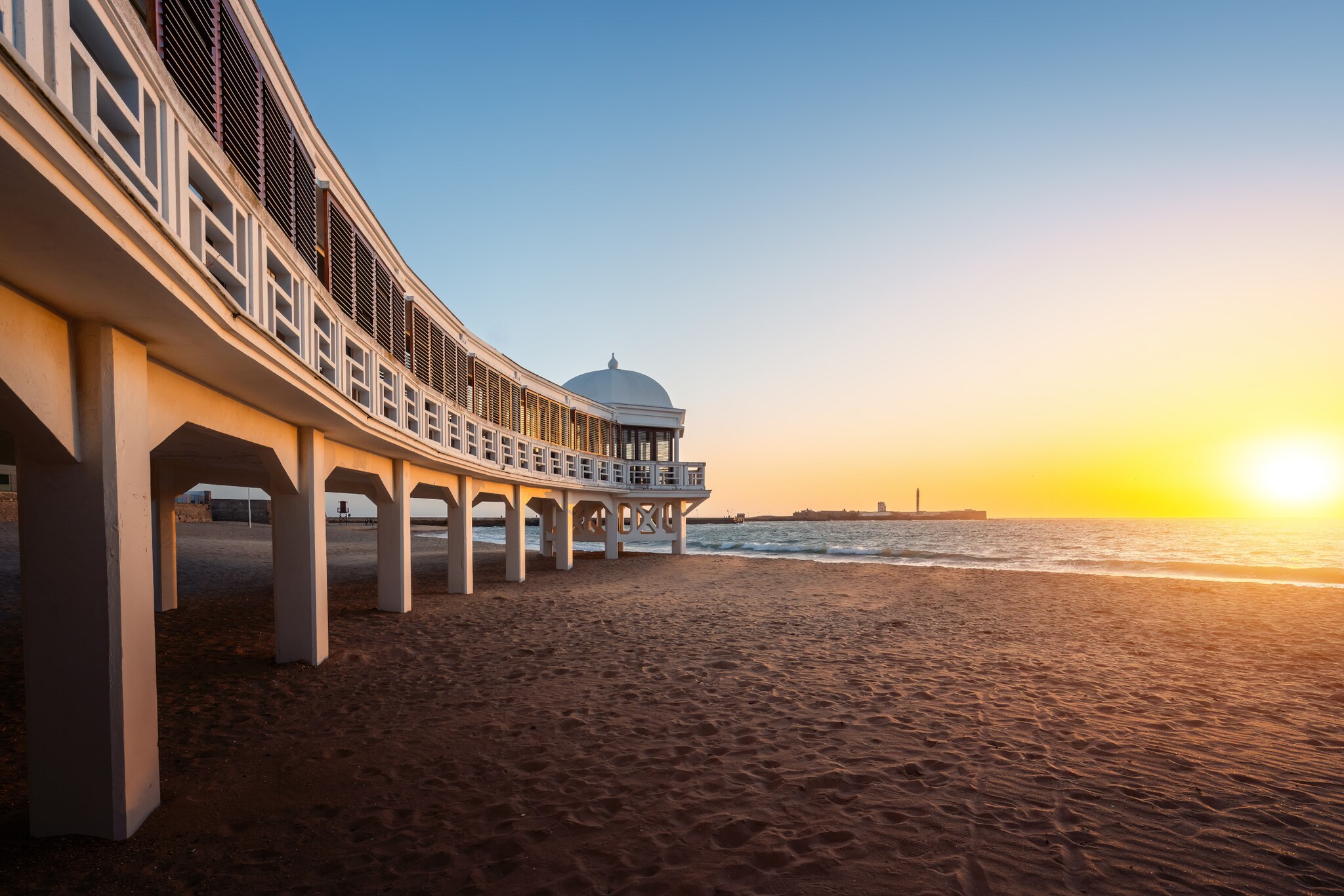 Strand bei Sonnenuntergang, am linken Bildrand ein weißes, langes Gebäude auf Stelzen.