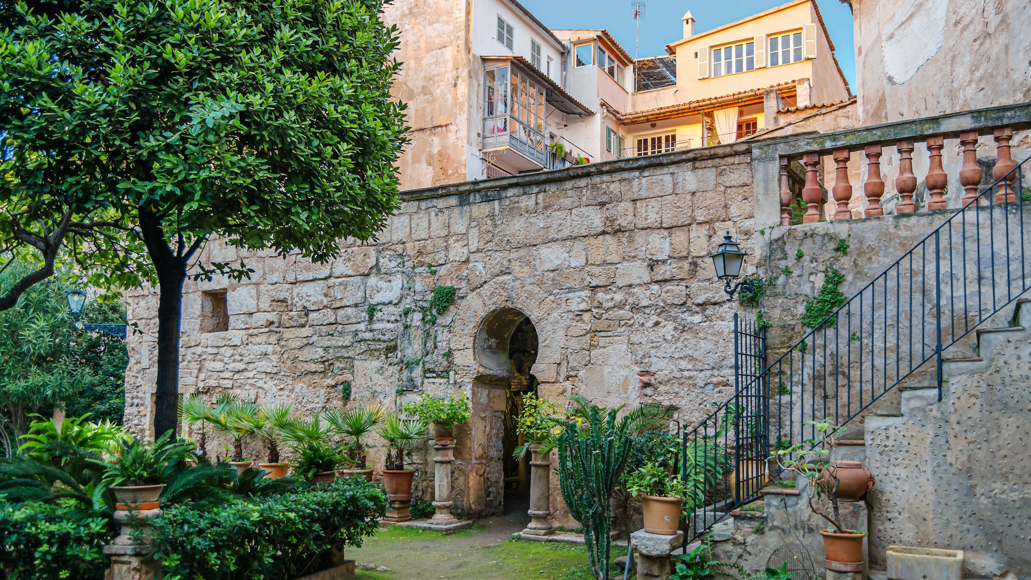 amex-magazin-geheimtipps-palma-innenhof-treppe Begrünter Innenhof mit Topfpflanzen vor historischem Mauerwerk mit einem Durchgang mit Hufeisenbogen.
