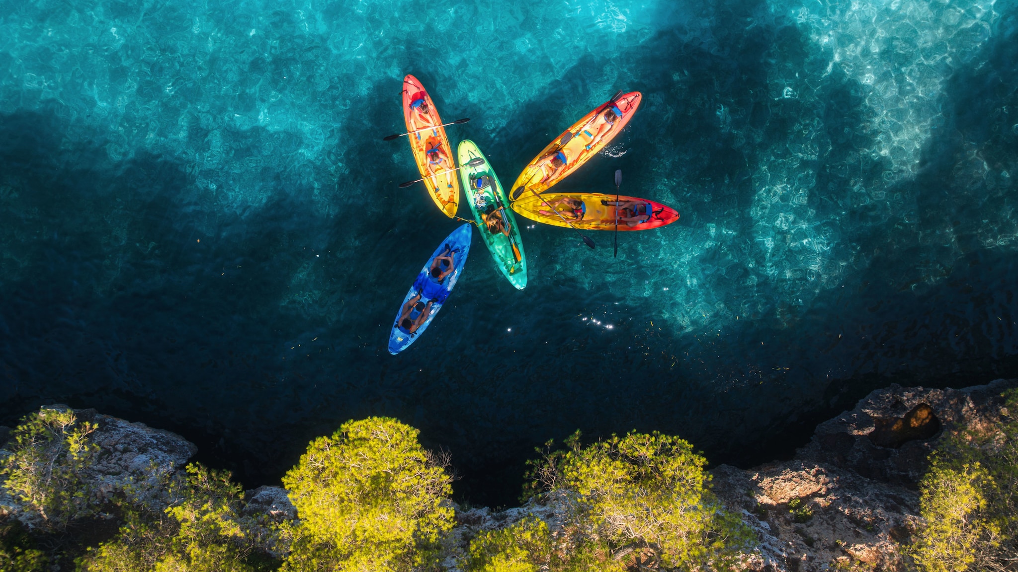 amex-magazin-geheimtipps-palma-kajaks Personen auf fünf Kajaks in unterschiedlichen Farben im türkisblauen Wasser an einer Felsküste aus der Vogelperspektive.
