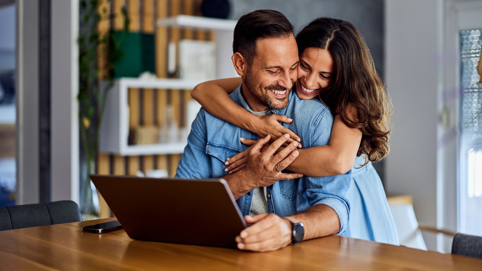 Eine Frau umarmt einen Mann, der an einem Schreibtisch sitzt.