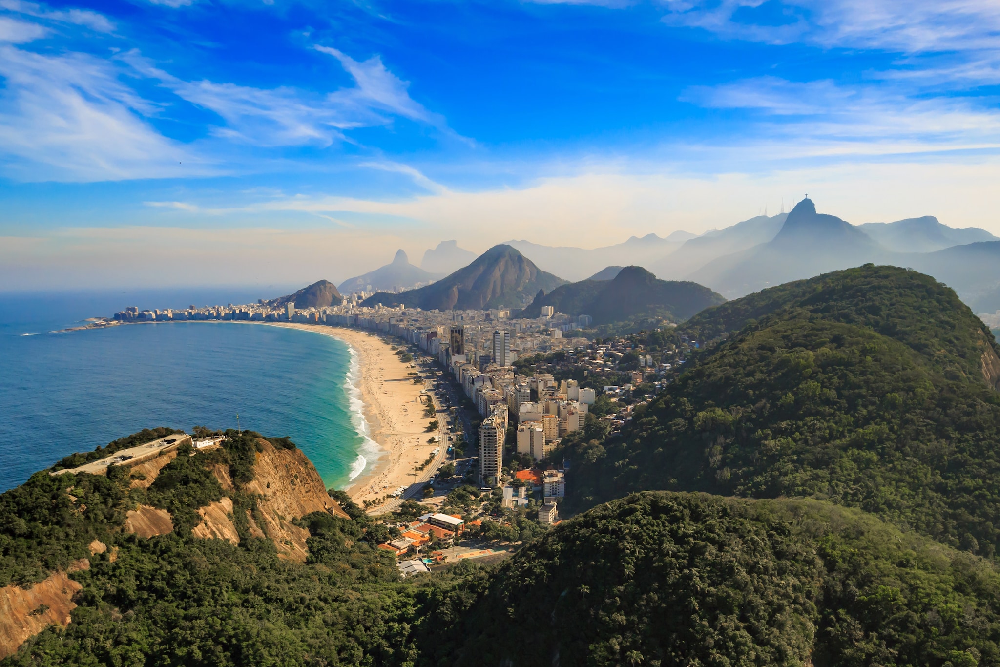 amex-magazin-brasilien-rundreise-meer-strand-stadt Blick von oben auf einen langen Sandstrand und eine große Stadt, die von Meer und grünen Hügeln umgeben sind.