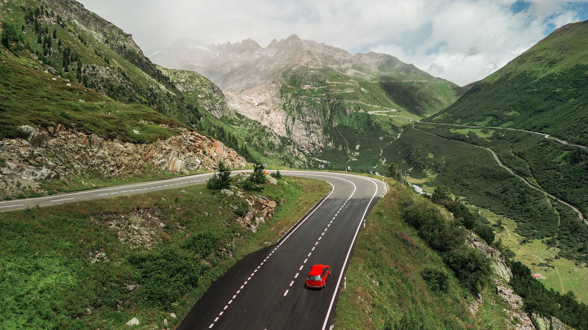 Ein rotes Auto fährt auf einer Straße, zwischen Bergen und grüner Landschaft.