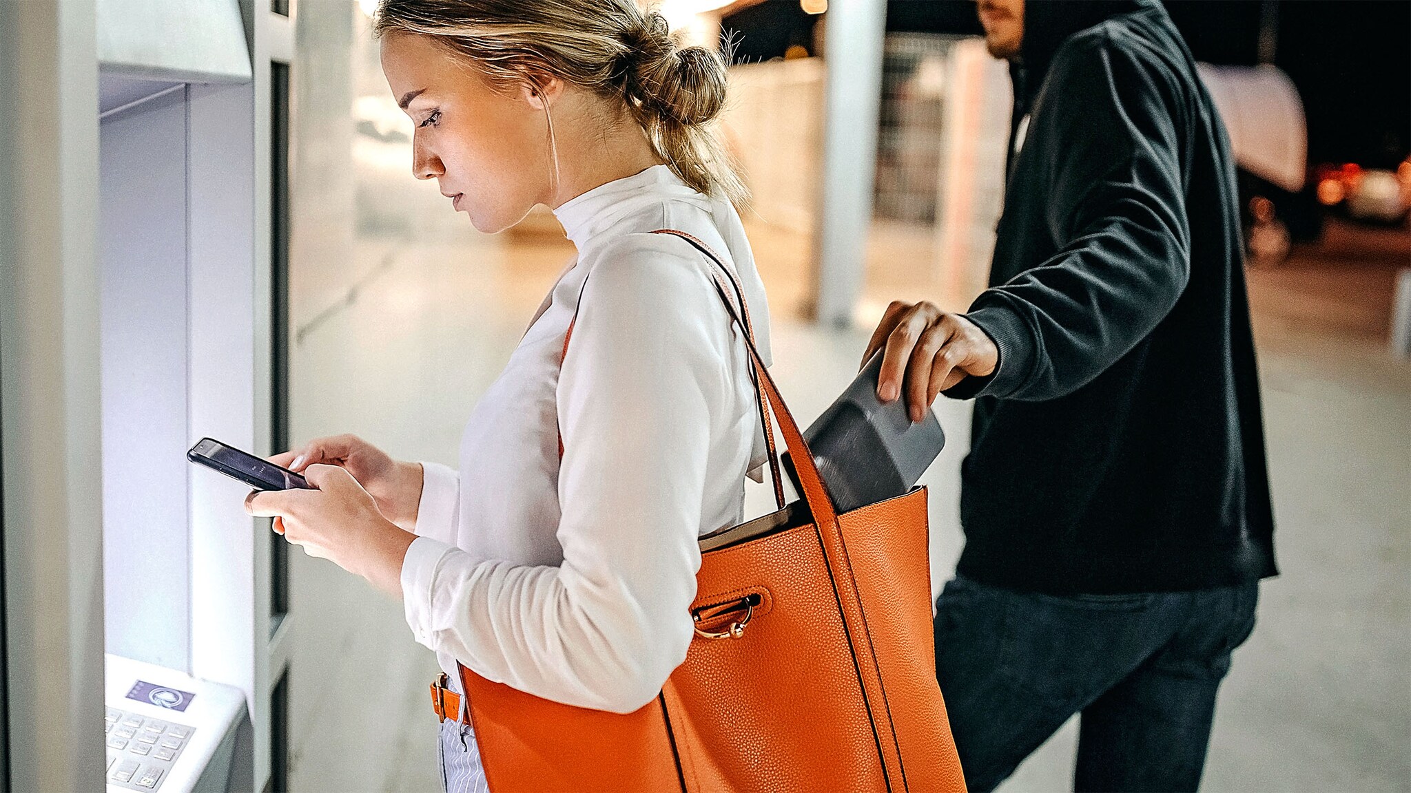 Eine junge Frau schaut auf ihr Handy an einem Geldautomaten, während ein Mann im schwarzen Kapuzenpullover ein Portemonnaie aus ihrer Handtasche entwendet in einer verlassenen Gegend bei Dunkelheit. 