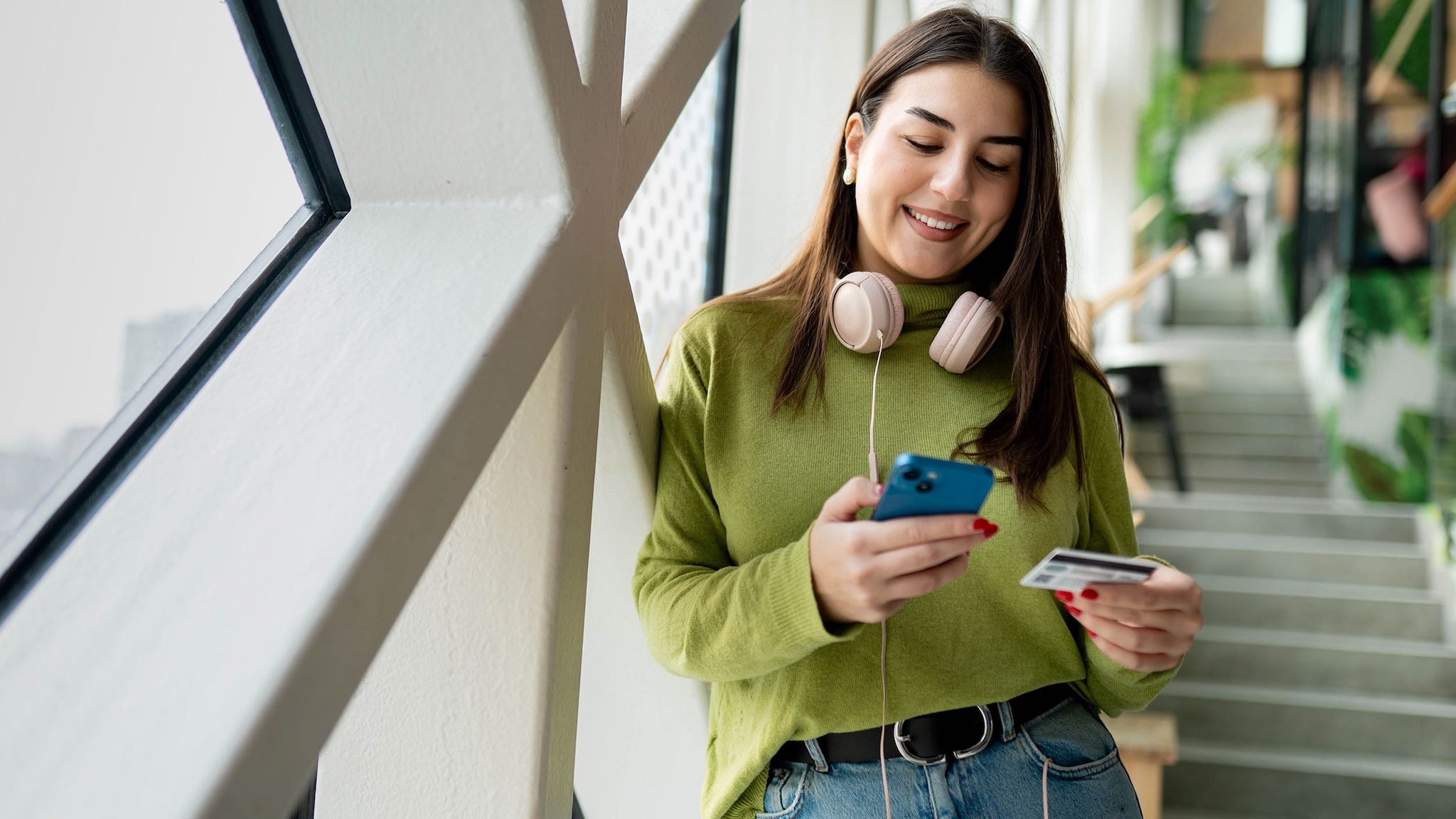 Eine junge Frau mit Kopfhörern um den Hals lehnt in einem Bürogang und schaut lächelnd auf ein Smartphone in ihrer Hand, während sie mit der anderen Hand eine Kreditkarte hält.