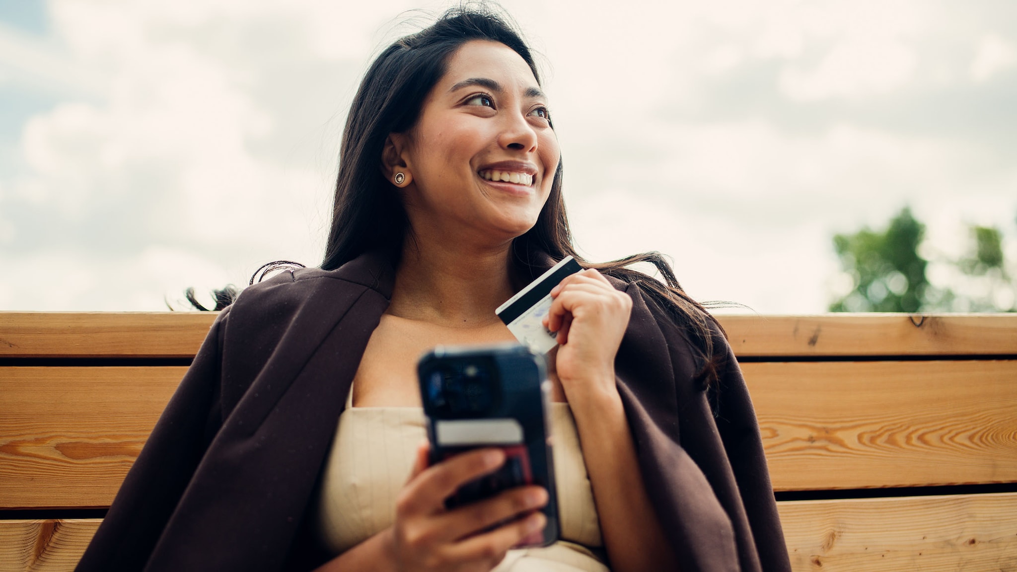 Eine elegant gekleidete Frau sitzt zufrieden lächelnd auf einer Bank und schaut in die Ferne, in ihren Händen hält sie ein Smartphone und eine Kreditkarte.