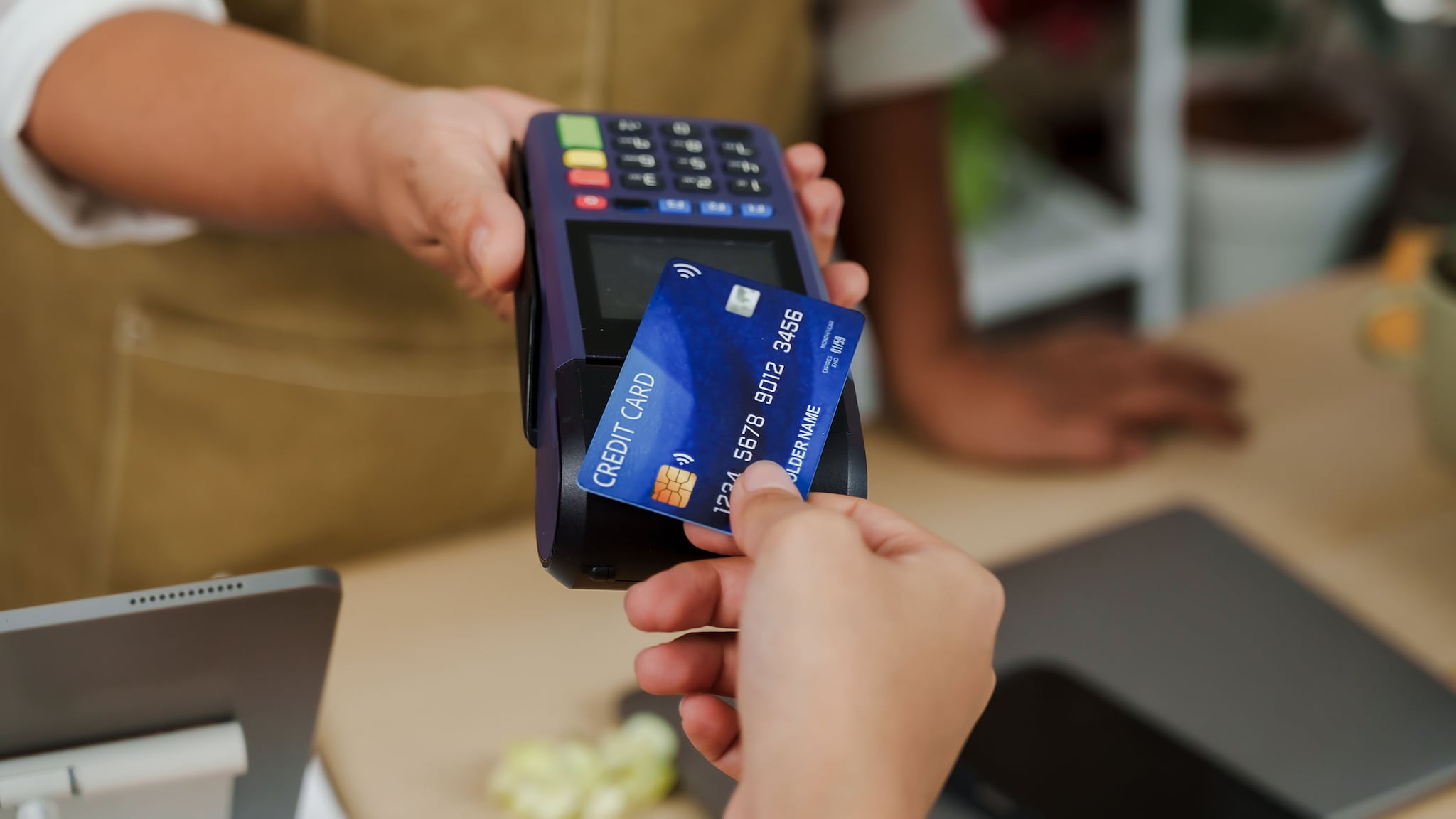 Kontaktlose Zahlung mit einer blauen Kreditkarte an einem Kartenlesegerät in der Hand einer Person hinter einem Tresen.