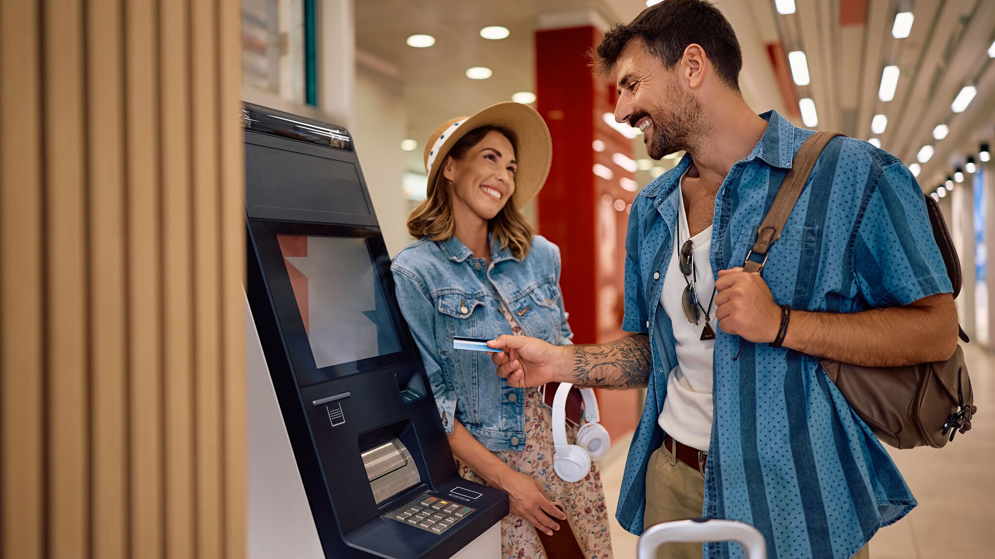 Eine Frau und ein Mann in Urlaubskleidung und mit Reisegepäck stehen gut gelaunt an einem Bargeldautomaten in einem Flughafenterminal, er hält eine Kreditkarte in der Hand.