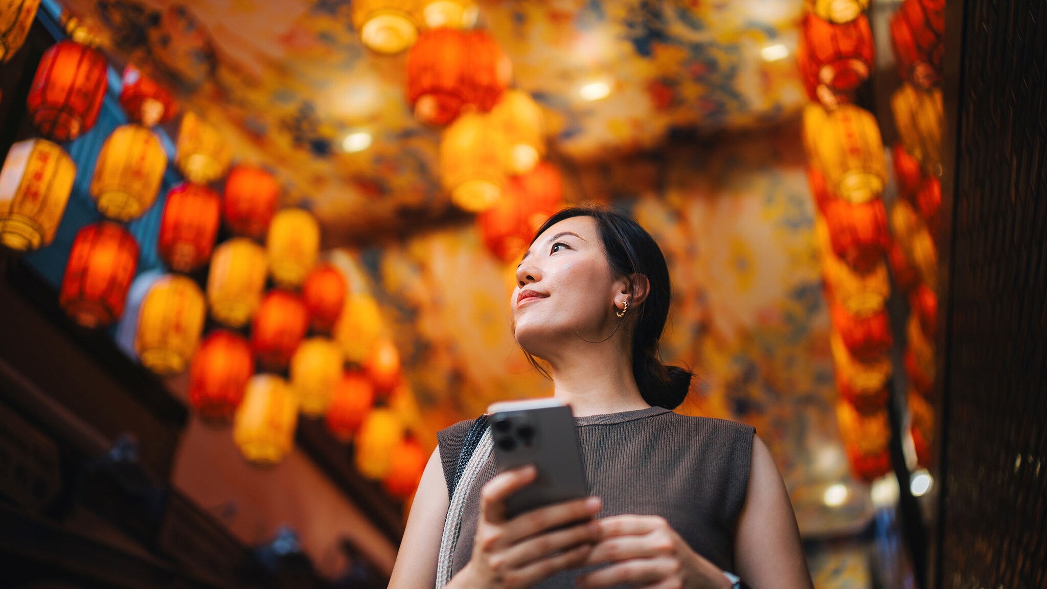 Porträt einer asiatischen Frau mit Smartphone in der Hand vor leuchtenden orangefarbenen Lampions in einer Straße aus der Untersicht.