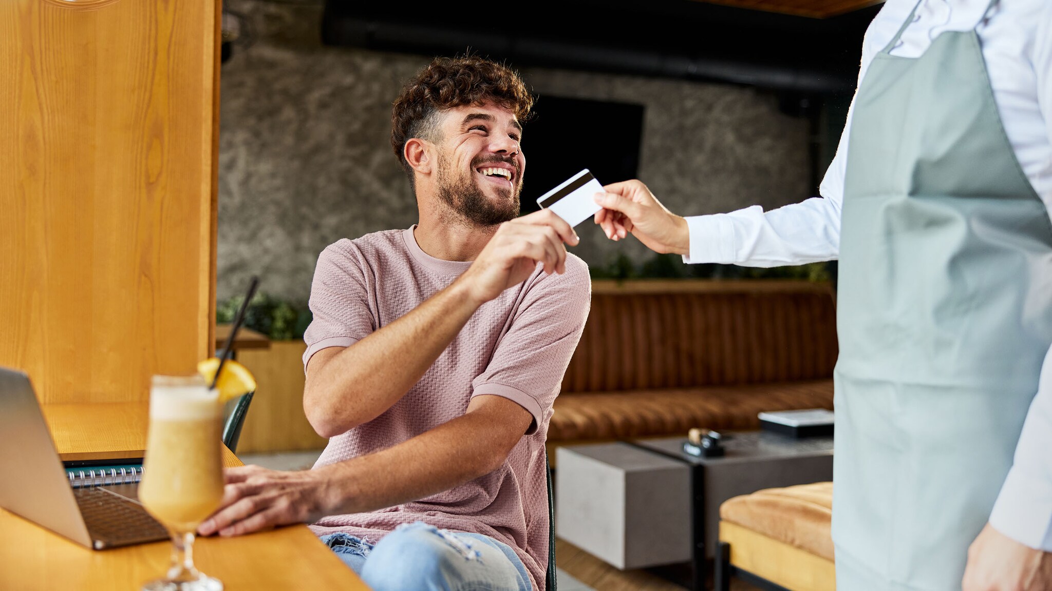 Ein Mann sitzt mit Laptop und Fruchtcocktail an einem Tisch in einer Lobby und reicht gut gelaunt einem Kellner eine Kreditkarte.