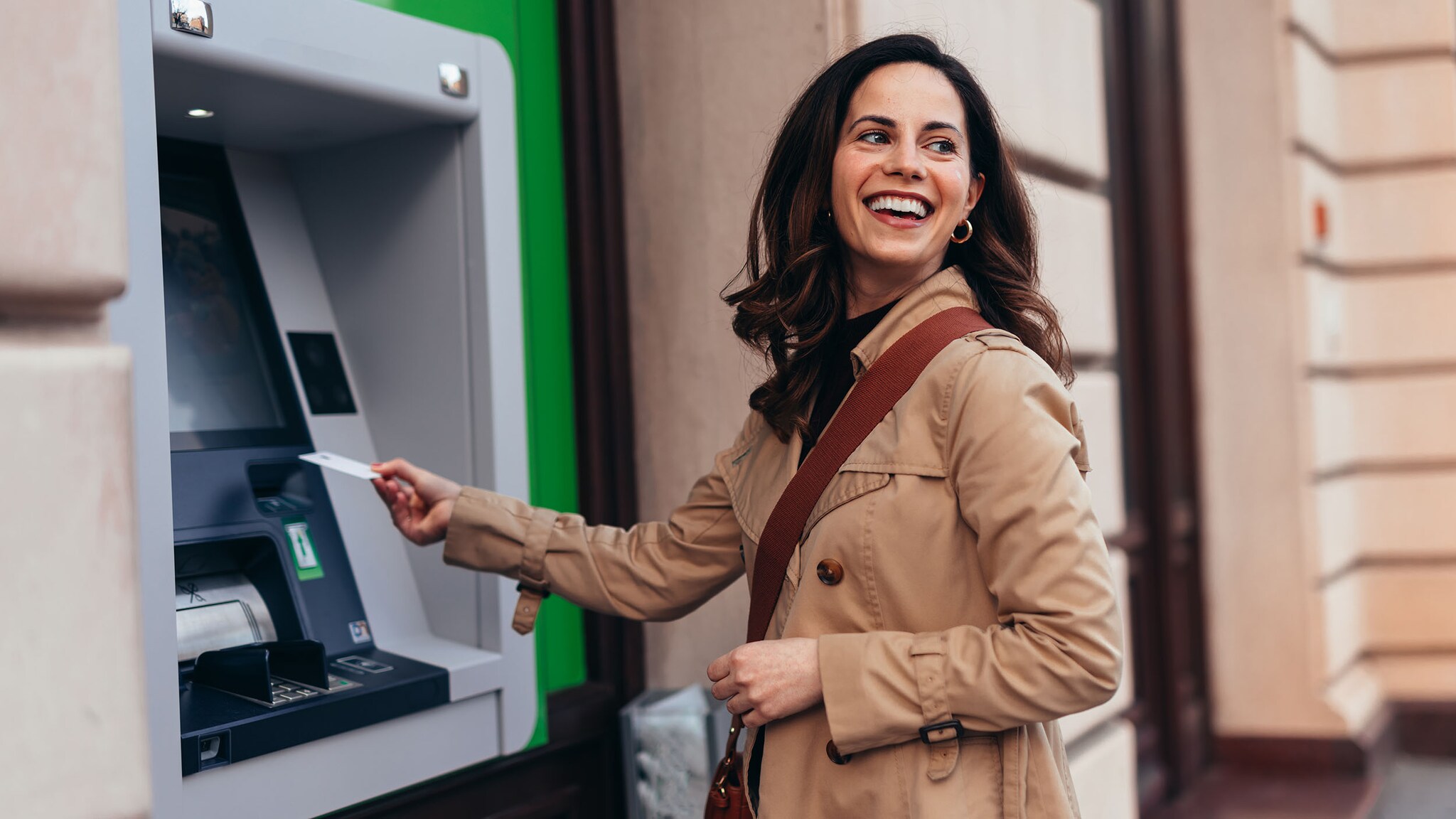 Eine lachende Frau im Trenchcoat steht mit einer Kreditkarte in ihrer Hand an einem Geldautomaten auf einer Straße.