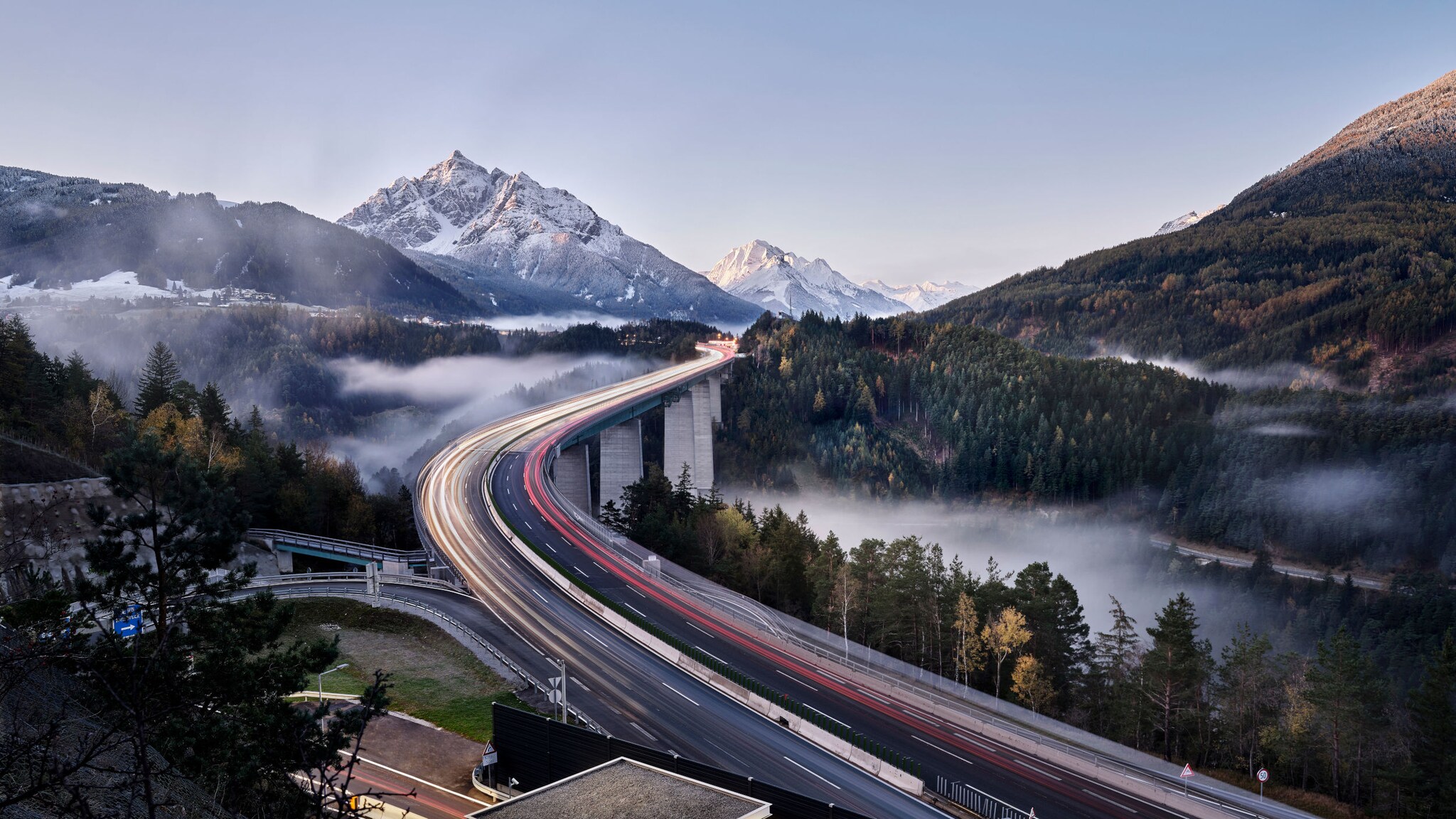 Landschaftspanorama mit Lichtstreifen auf einer geschwungenen Autobahnbrücke in einem hügeligen Waldgebiet vor schneebedeckten Bergen bei Abenddämmerung.