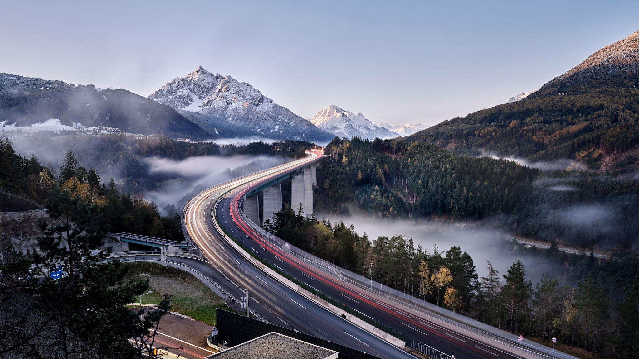 Landschaftspanorama mit Lichtstreifen auf einer geschwungenen Autobahnbrücke in einem hügeligen Waldgebiet vor schneebedeckten Bergen bei Abenddämmerung. Landschaftspanorama mit Lichtstreifen auf einer geschwungenen Autobahnbrücke in einem hügeligen Waldgebiet vor schneebedeckten Bergen bei Abenddämmerung.
