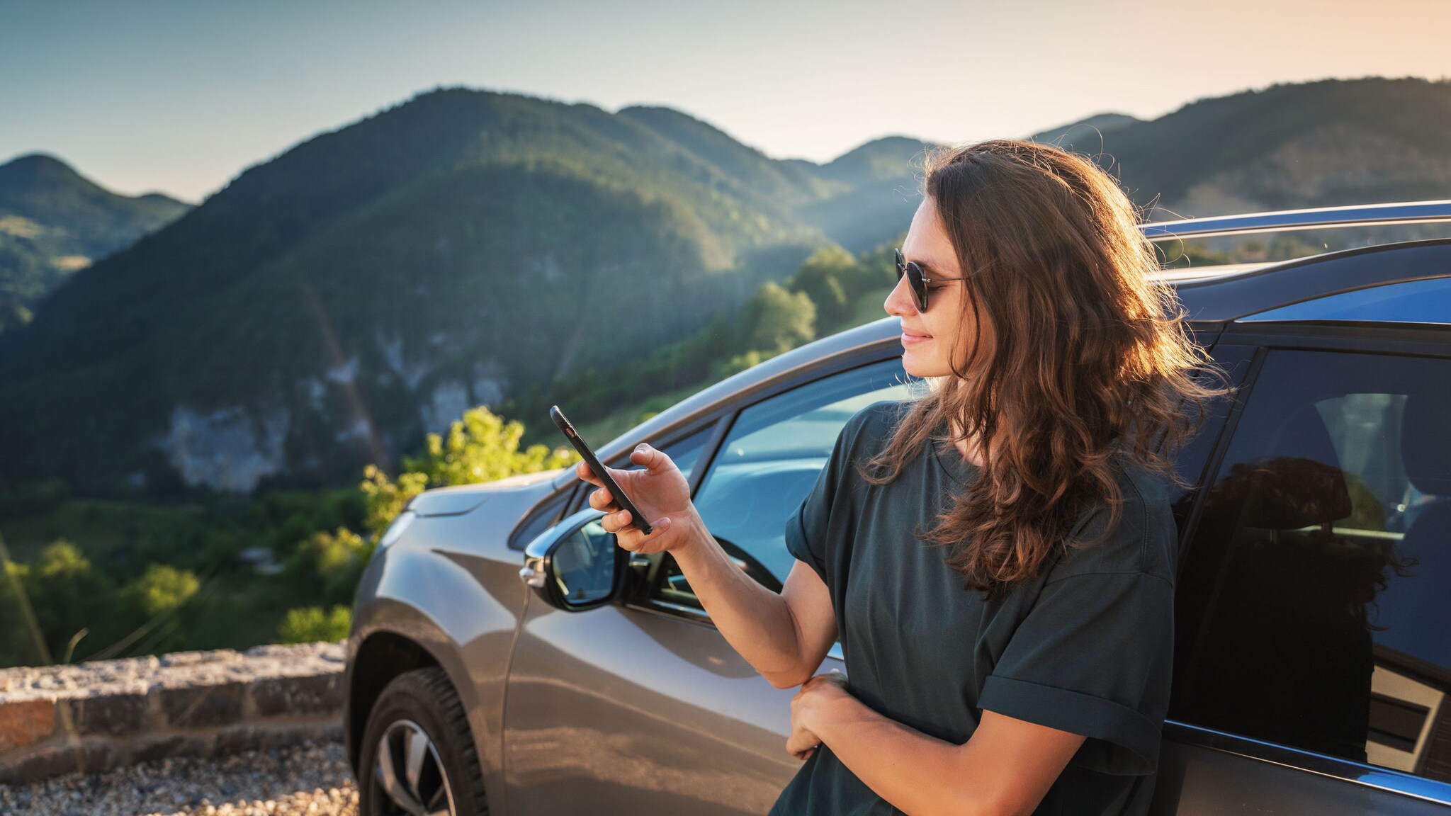 Eine junge Frau mit Sonnenbrille lehnt an einem geparkten Auto und schaut auf ihr Smartphone vor einer bewaldeten Berglandschaft bei Sonnenschein.