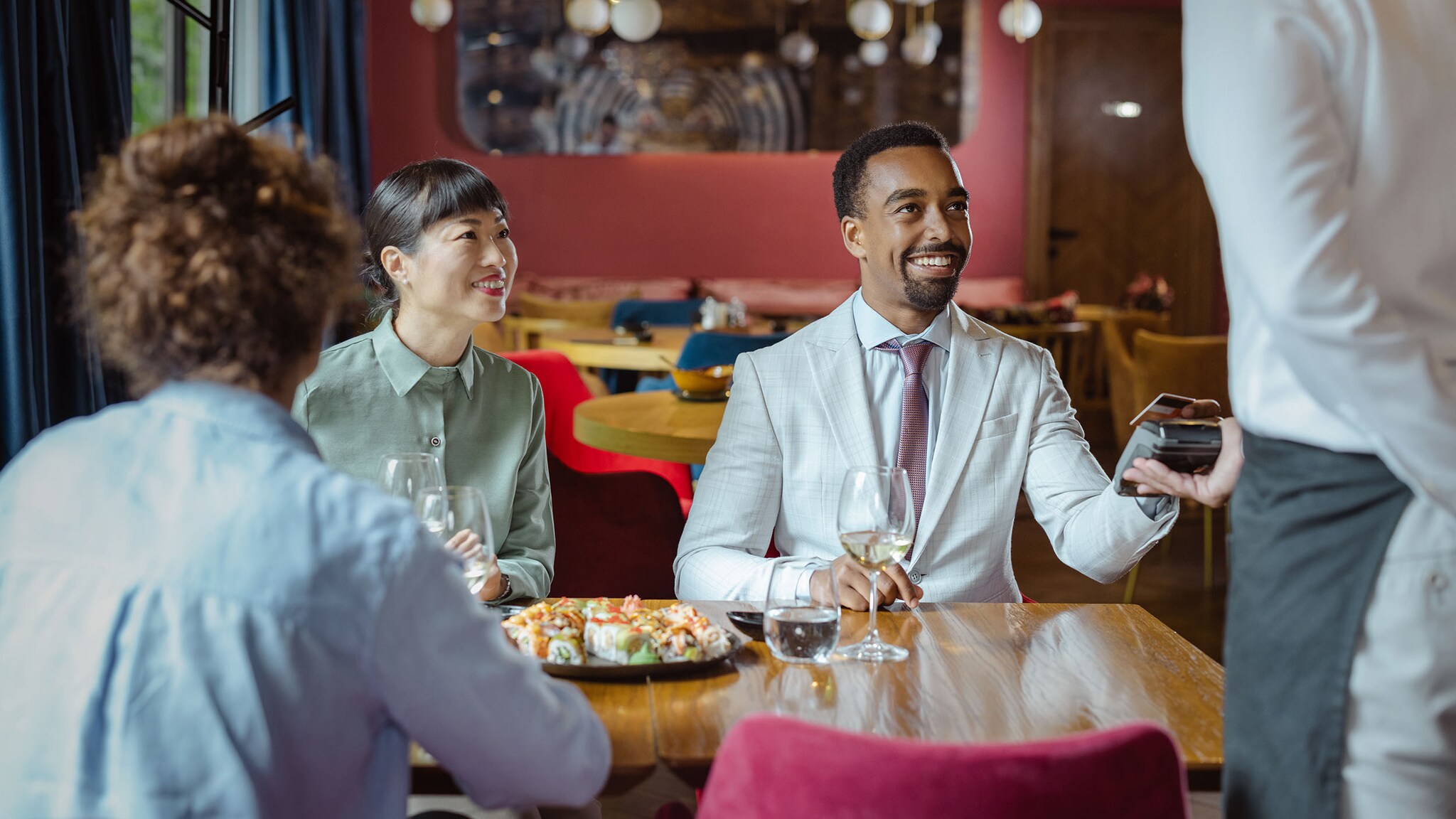 Ein lächelnder Mann an einem Tisch mit drei weiteren Personen bezahlt kontaktlos mit einer Kreditkarte an einem Kartenlesegerät in der Hand eines Kellners in einem Restaurant.