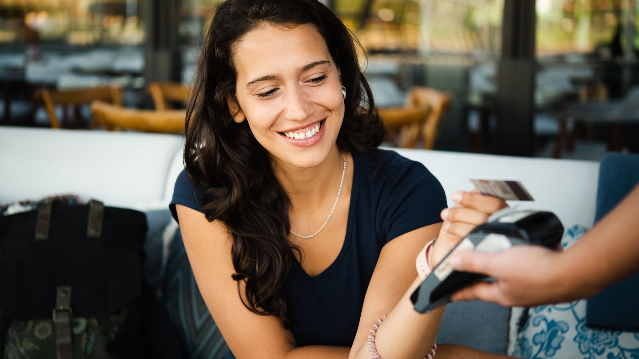 Close-up einer lächelnden Frau in Sommerkleidung, die auf der Terrasse eines Restaurants sitzt und kontaktlos mit Kreditkarte an einem Kartenlesegerät in der Hand einer Person zahlt.
