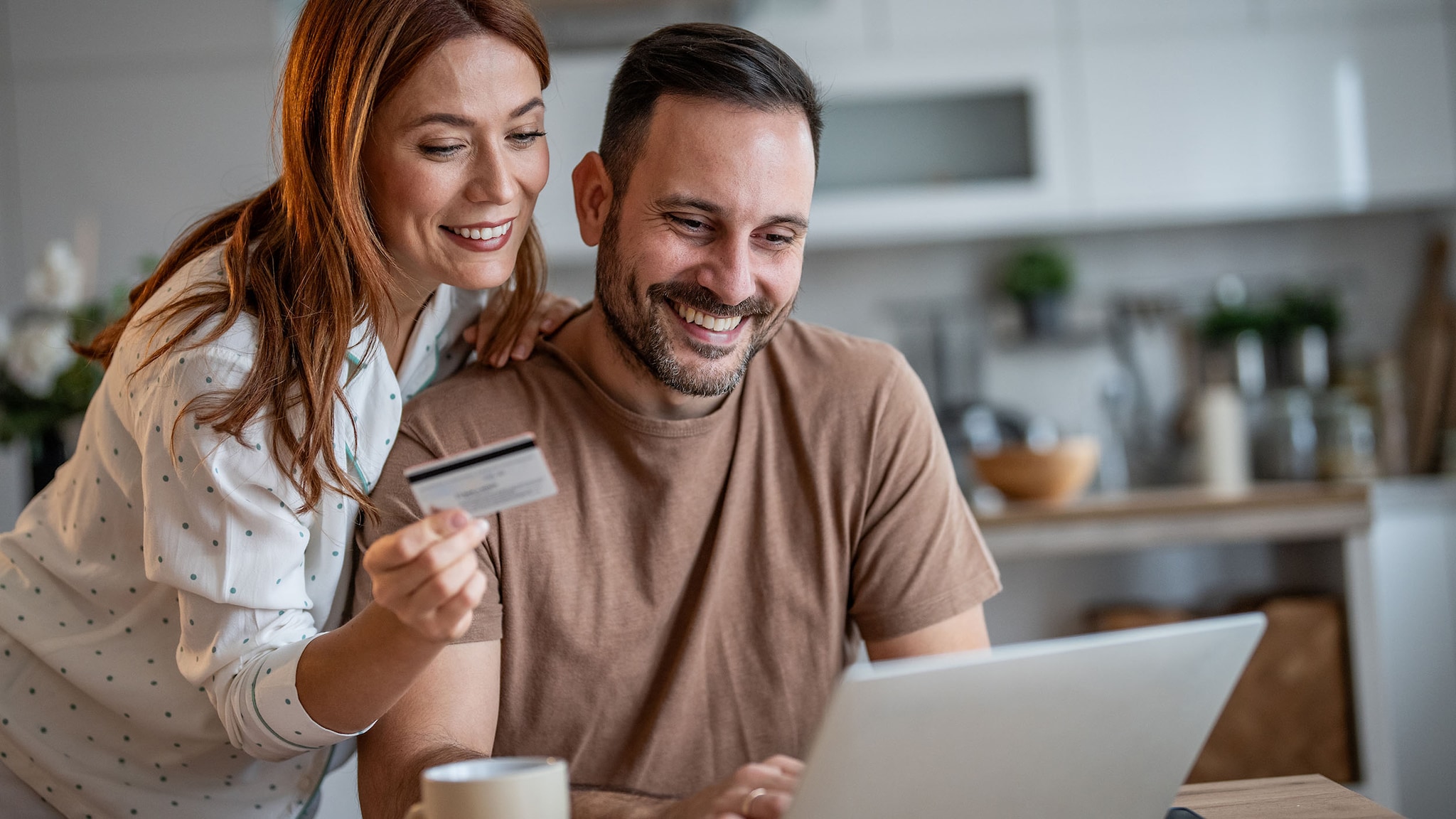 Ein Mann und eine Frau mit Kreditkarte in der Hand sitzen lächelnd vor einem Laptop auf einem Tisch in einer Küche.