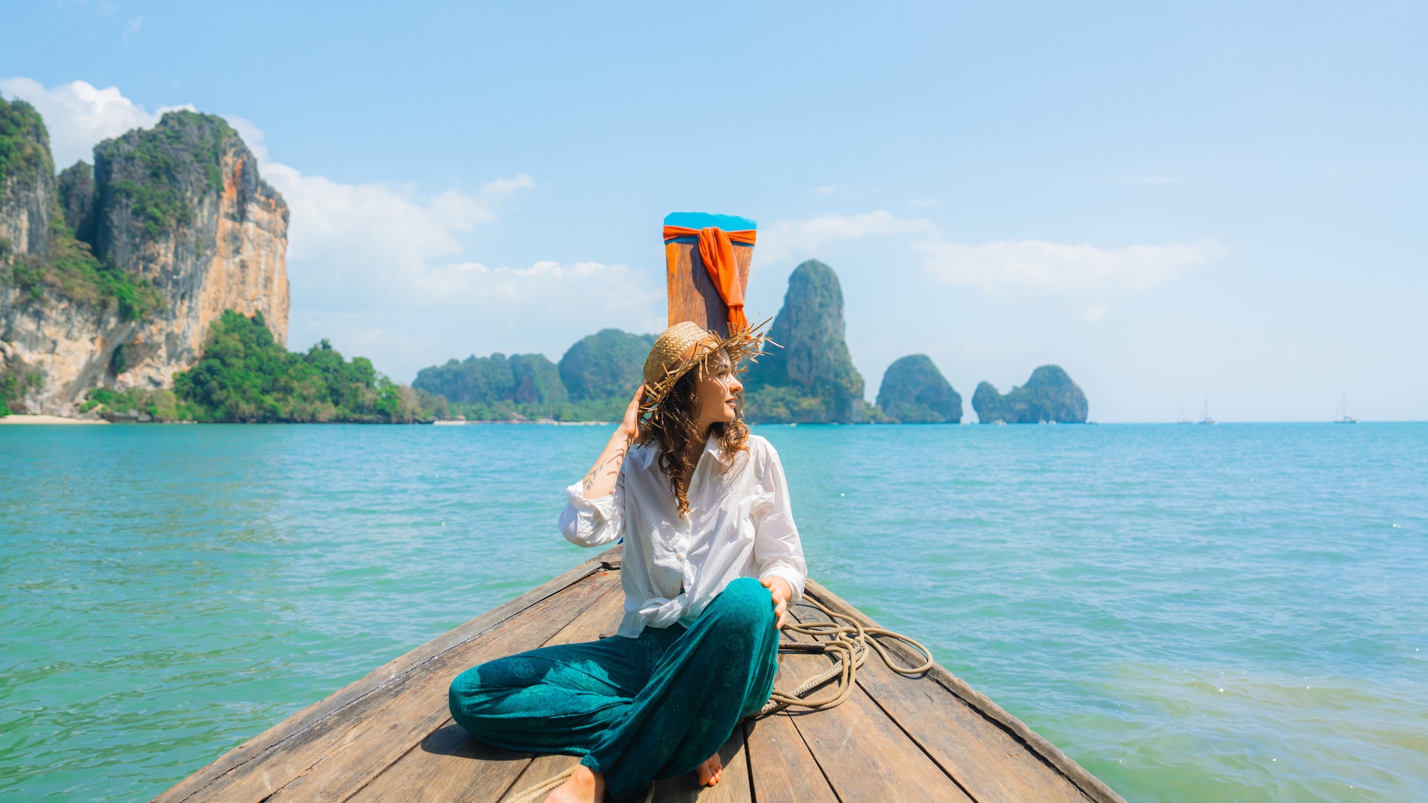 amex-magazin-geheimtipps-phuket-frau-holzboot Eine Frau mit Sonnenhut auf einem Holzboot auf türkisblauem Meer, im Hintergrund grün bewachsene Felsen.