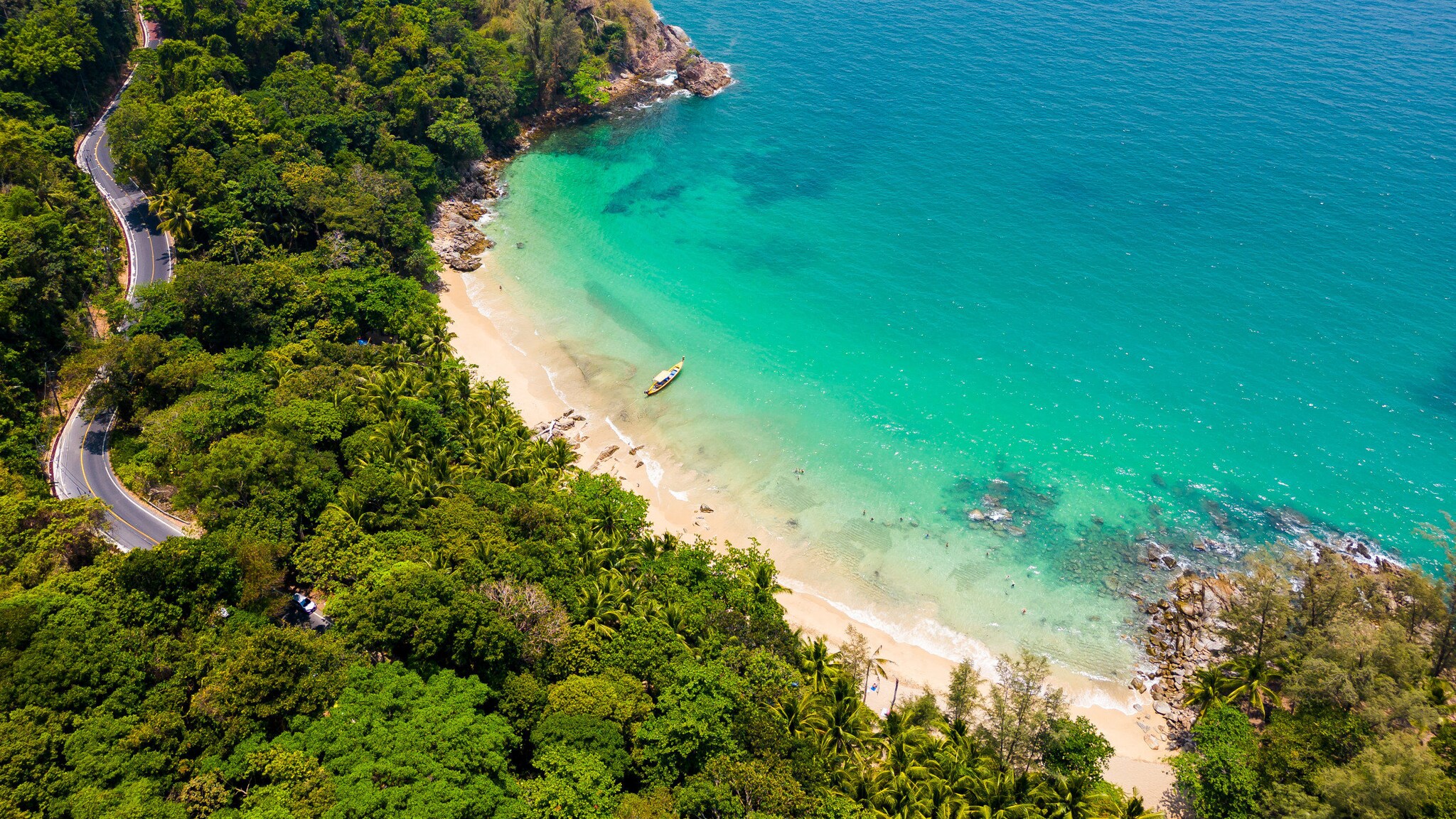 Luftaufnahme eines Strandes mit türkisblauem Wasser, der von dichter grüner Vegetation umgeben ist, durch die eine Straße führt.