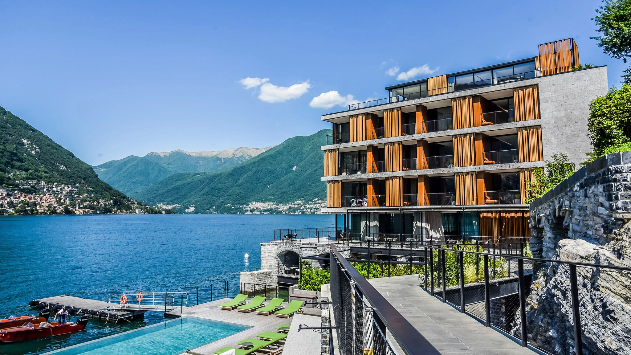 kastenförmiges, dreistöckiges Hotel mit Terrasse an einem See vor mediterranem Bergpanorama.