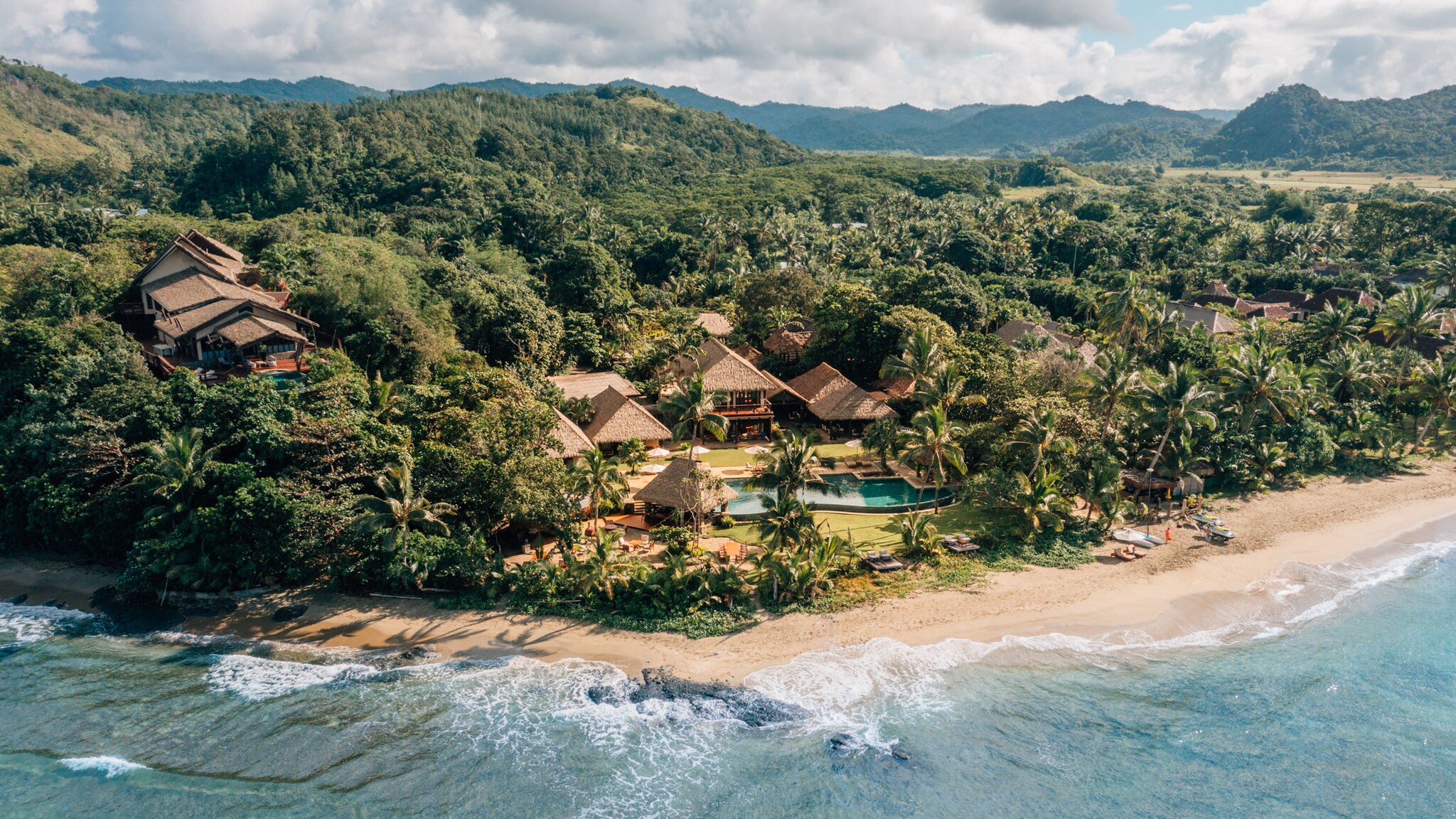 Luxus-Hotel-Resort mit strohbedeckten Bungalows mit Strohdächern und Pool, umgeben von Urwald an einem Sandstrand an einer Meeresküste.