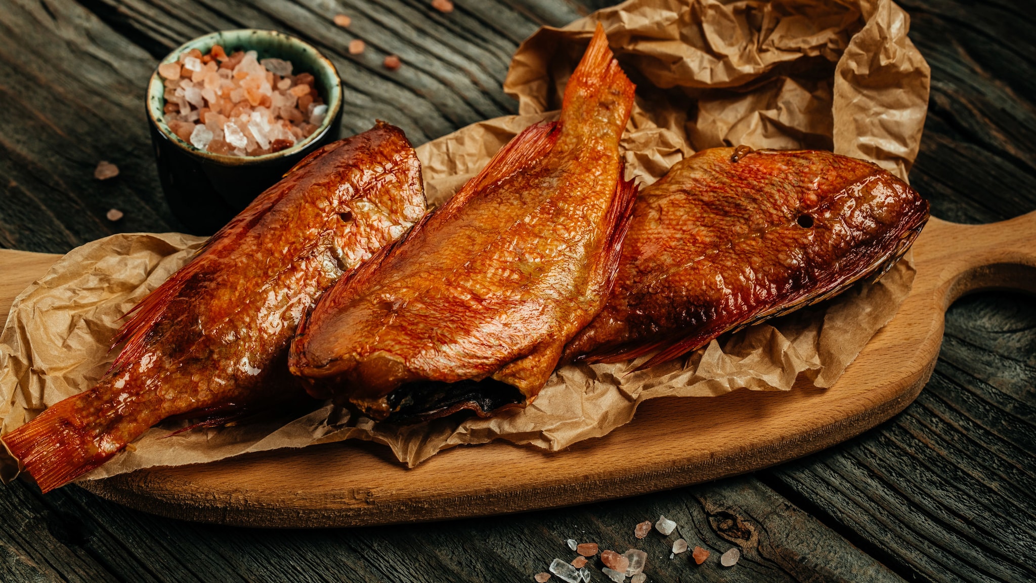 Drei geräucherte Fischstücke mit goldbrauner Kruste liegen angerichtet auf einem Holzbrett. Drei geräucherte Fischstücke mit goldbrauner Kruste liegen angerichtet auf einem Holzbrett.