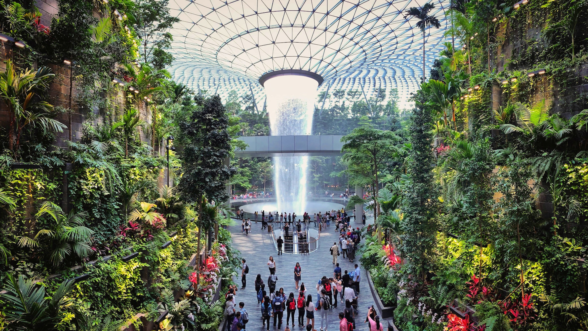 Personen in einem Flughafenterminal mit vertikalem tropischem Garten und Indoor-Wasserfall, der aus einer Glaskuppel fällt.