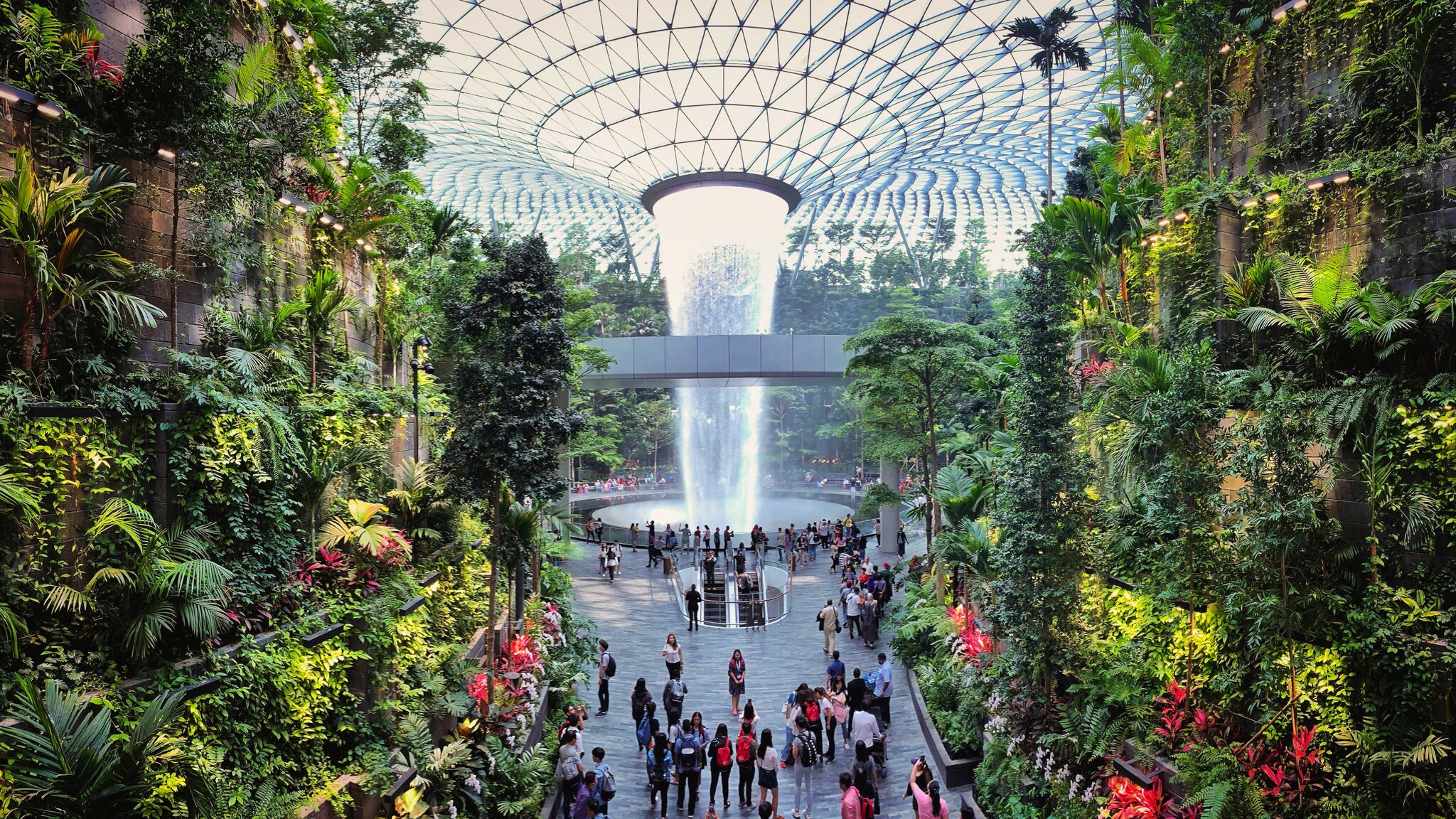 amex-magazin-singapur-urlaub-tropischer-garten Personen in einem Flughafenterminal mit vertikalem tropischem Garten und Indoor-Wasserfall, der aus einer Glaskuppel fällt.
